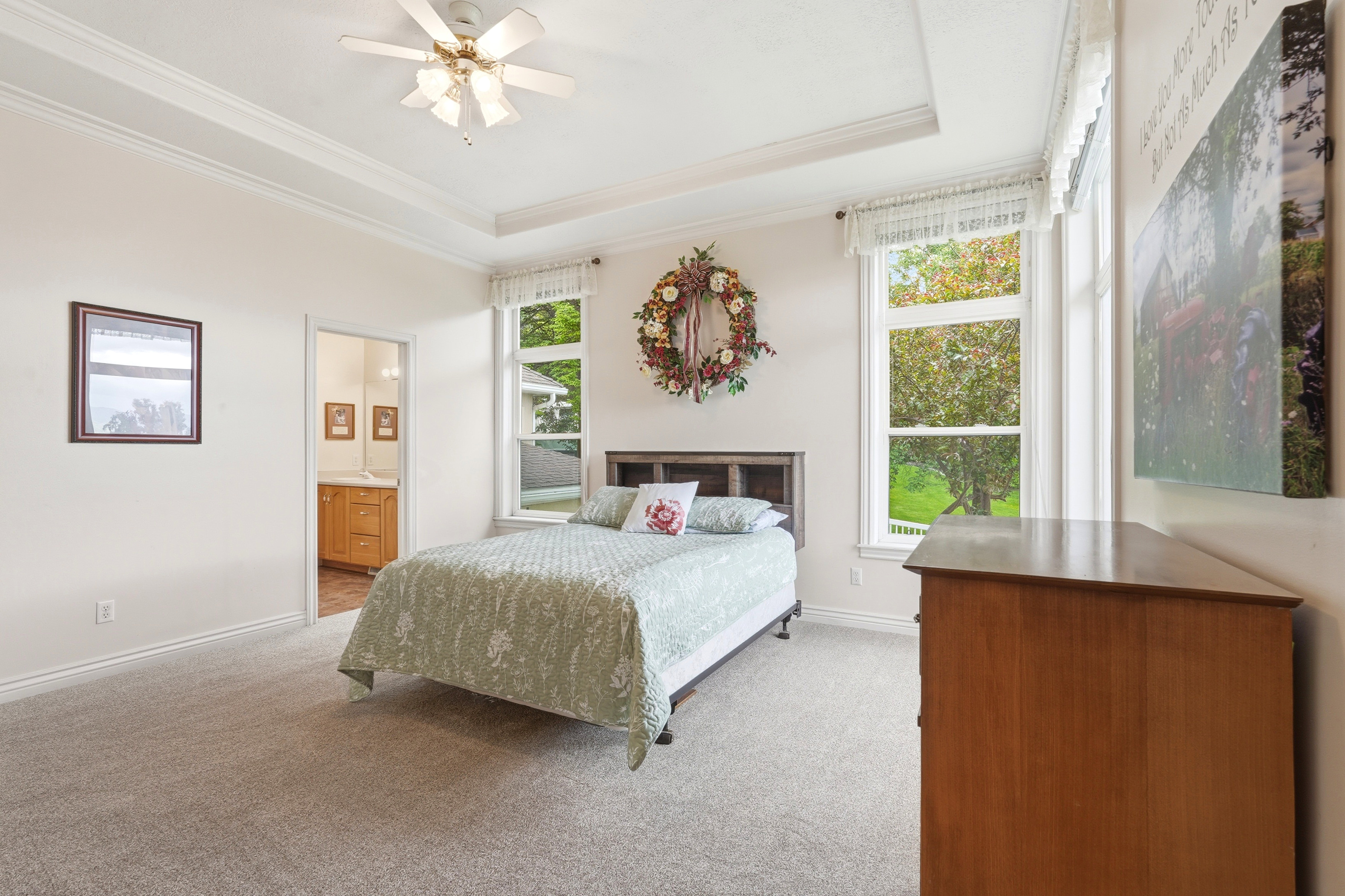 Carpeted room featuring a tray ceiling with a ceiling fan, two large windows with white trim, and a doorway opening to a bathroom with a wood-finish vanity and mirror
