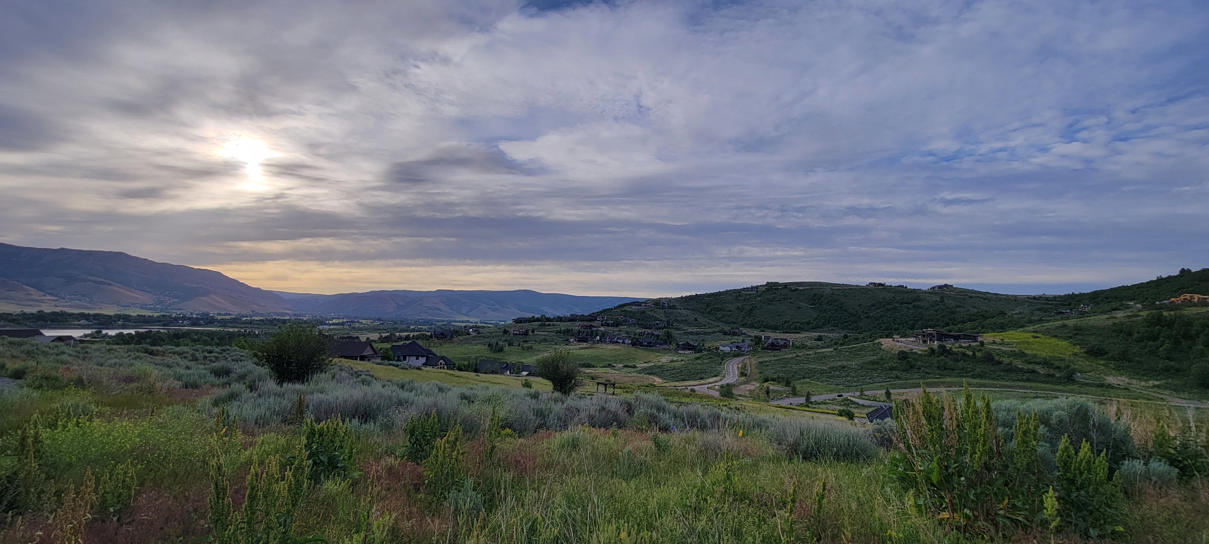 Expansive mountain landscape featuring varied topography, distant residential structures, and a body of water under a dramatic sky