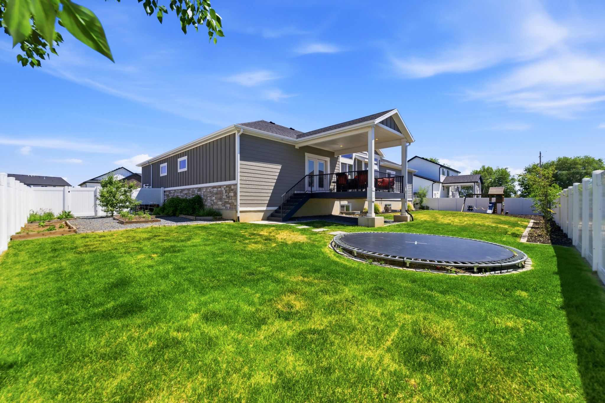 Spacious backyard featuring a covered deck, extensive green lawn, white privacy fencing, and garden beds.