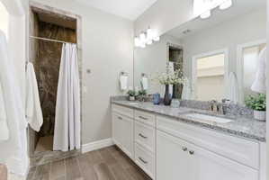 Bathroom featuring a dual vanity with granite-style countertops, wood-finish tile flooring, a walk-in shower with stone-look tile surround, and a large wall-mounted mirror