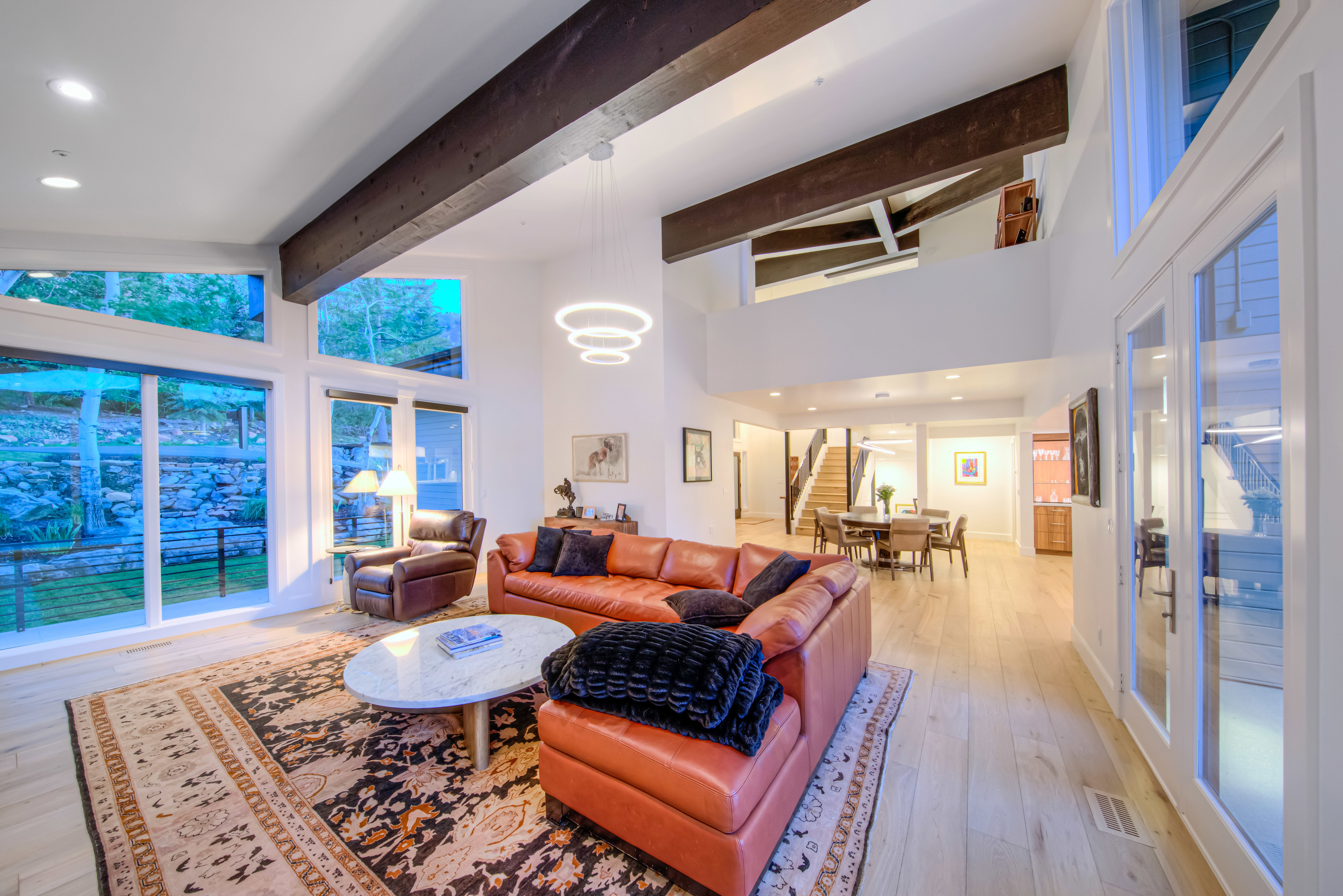 Vaulted ceilings with exposed wood beams define this expansive room, featuring large windows, wood-finish flooring, and a contemporary chandelier