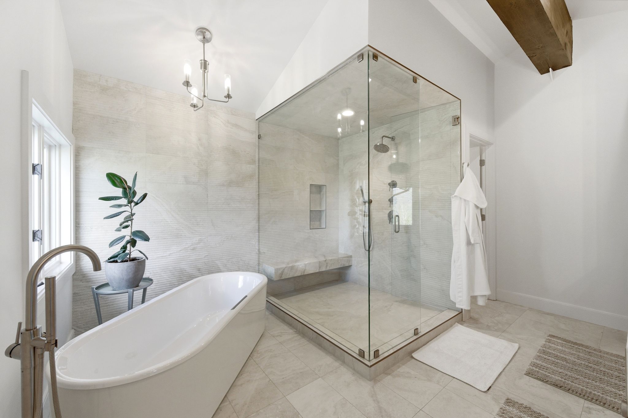 Spacious bathroom featuring a freestanding soaking tub, a large glass-enclosed shower with a built-in bench, light-toned tile flooring, and a exposed wood beam accent