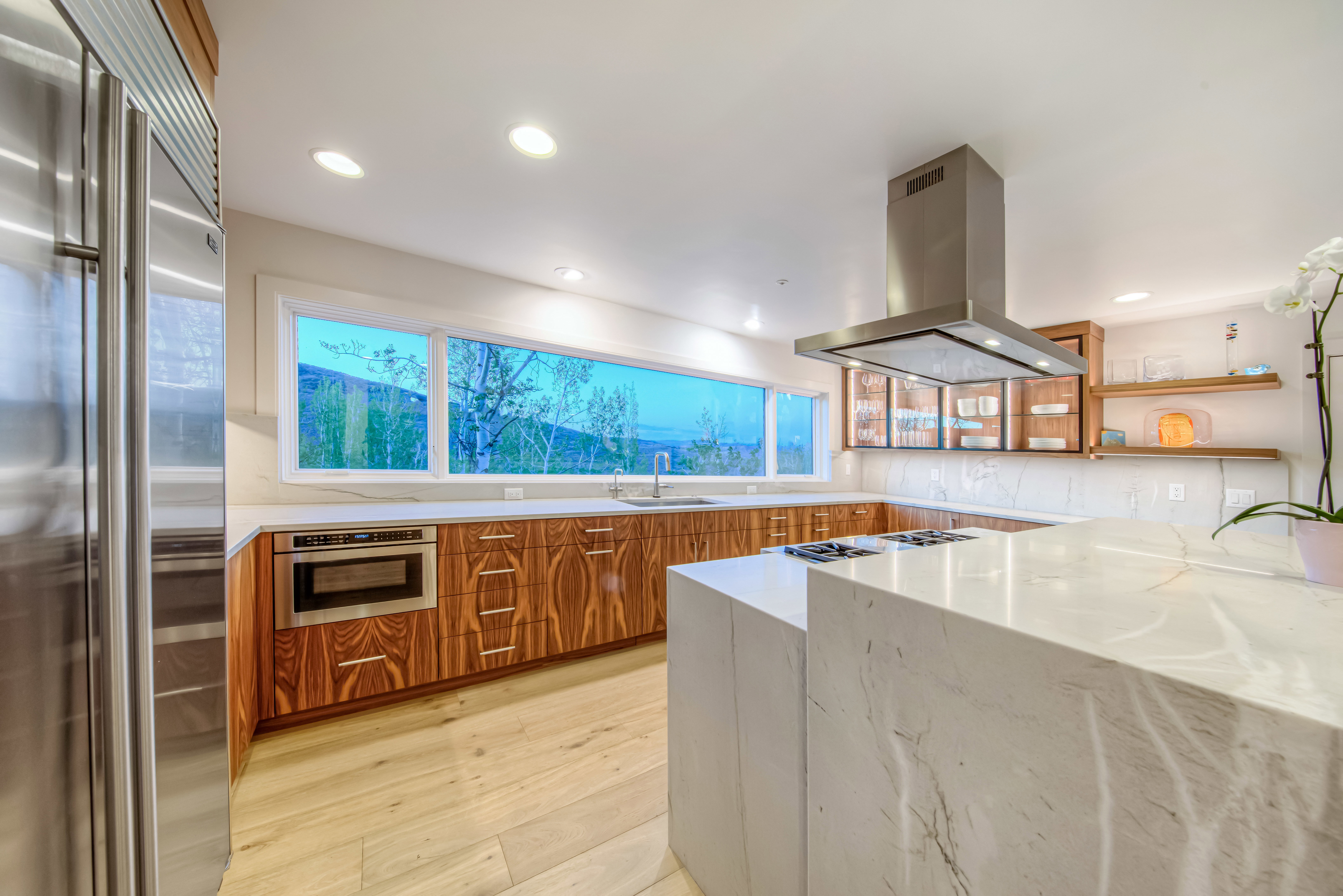 Modern kitchen featuring a large waterfall-edge island with an integrated cooktop, stainless steel appliances, extensive wood-finish cabinetry, a wide horizontal window, and recessed ceiling lighting