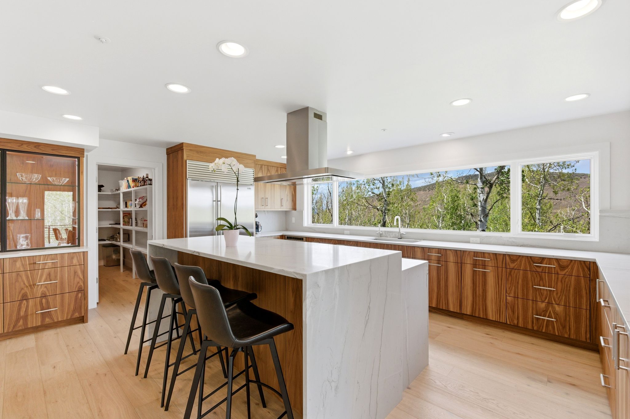 Kitchen featuring a substantial island with stone countertop, extensive wood-finish cabinetry, a stainless steel range hood, recessed lighting, and a panoramic window overlooking tree-covered hills