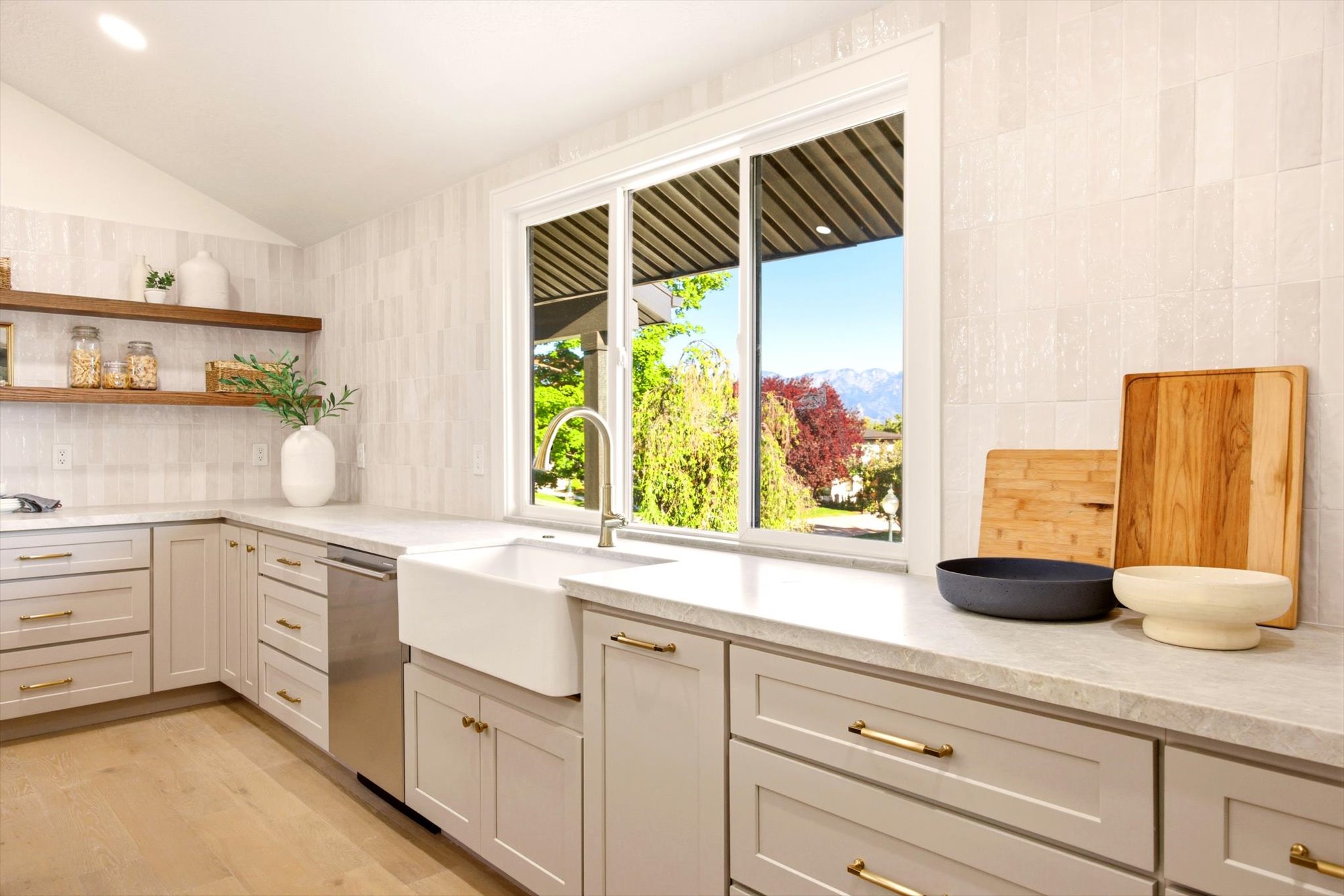 Kitchen featuring a large window, farmhouse sink with a gooseneck faucet, light grey cabinetry with gold-tone hardware, wood-finish flooring, and open wood shelving
