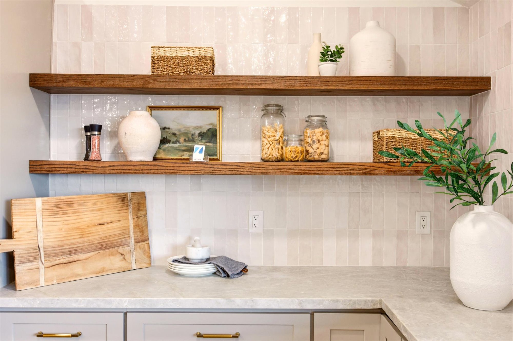 Kitchen featuring open wood shelving, light-toned vertical tile backsplash, light grey countertops, and light grey base cabinetry with brass hardware