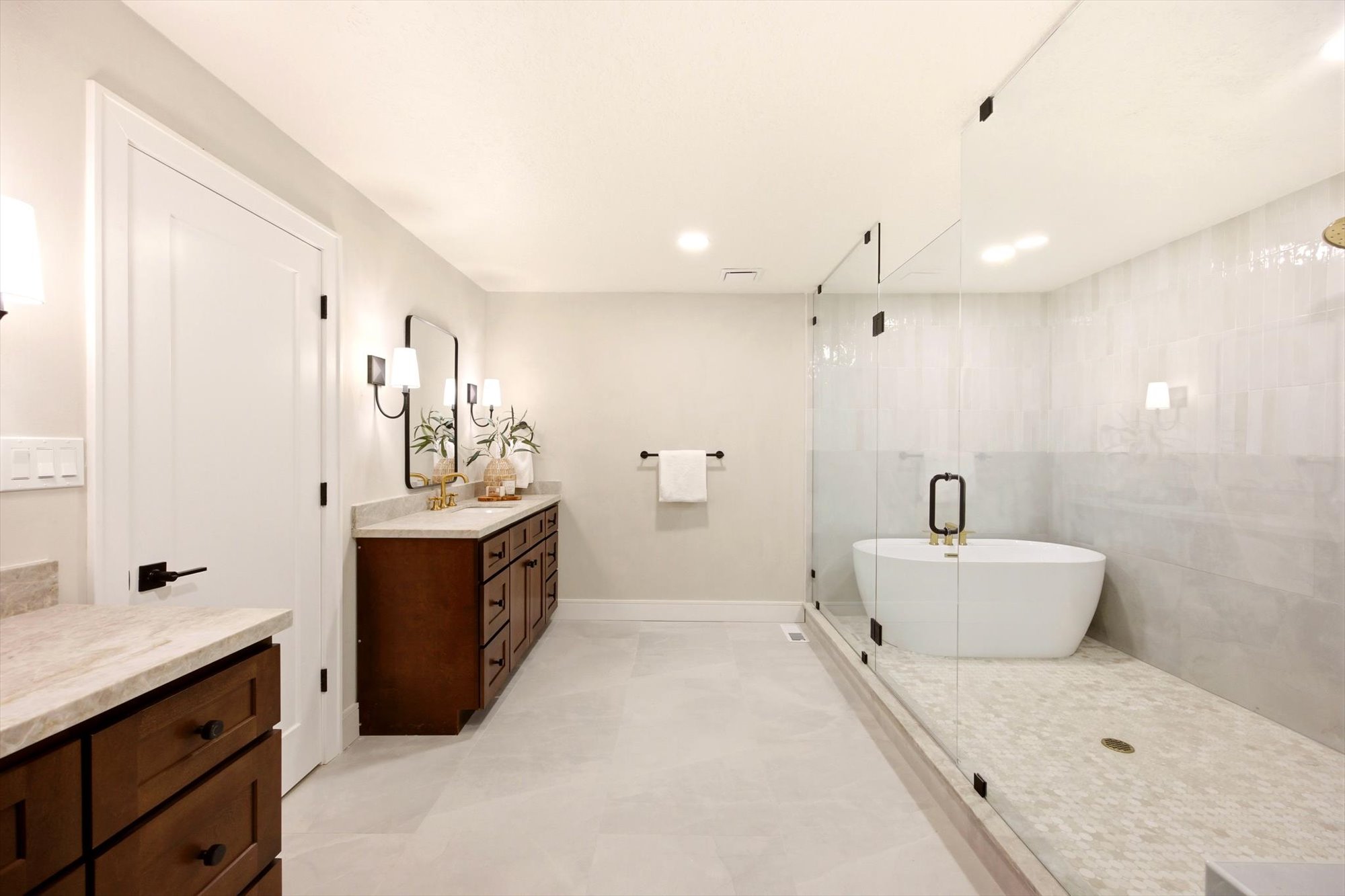 Spacious bathroom featuring a frameless glass shower enclosure with a freestanding soaking tub, dual vanity with stone countertops, dark wood-finish cabinetry, and contemporary wall sconces