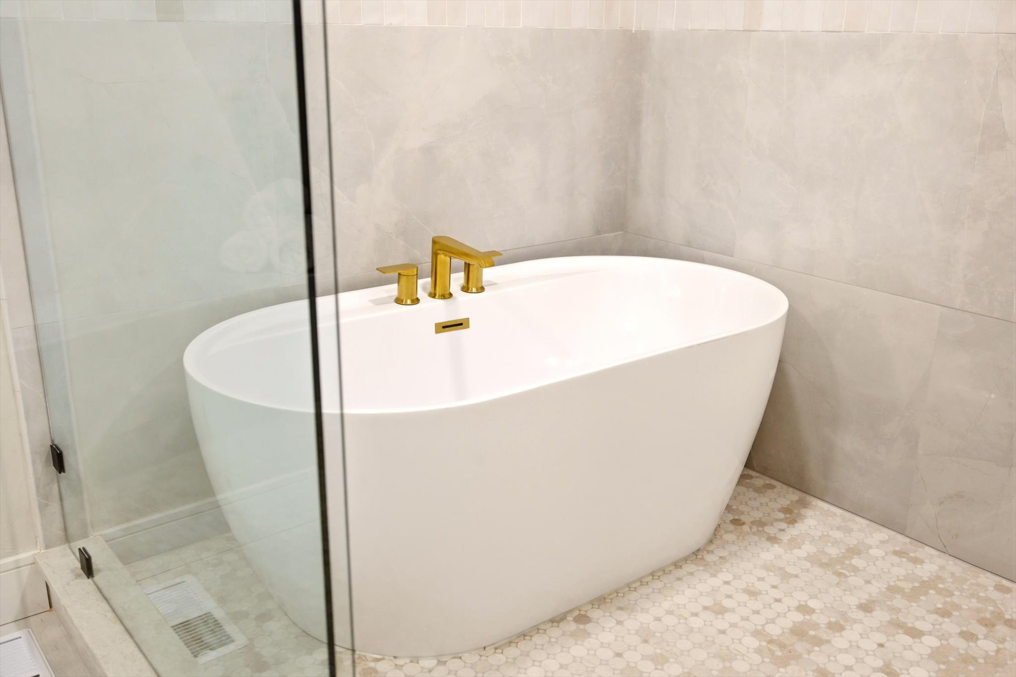 Freestanding oval soaking tub with gold-finish fixtures, complemented by light-toned wall tiling and hexagonal floor tiling