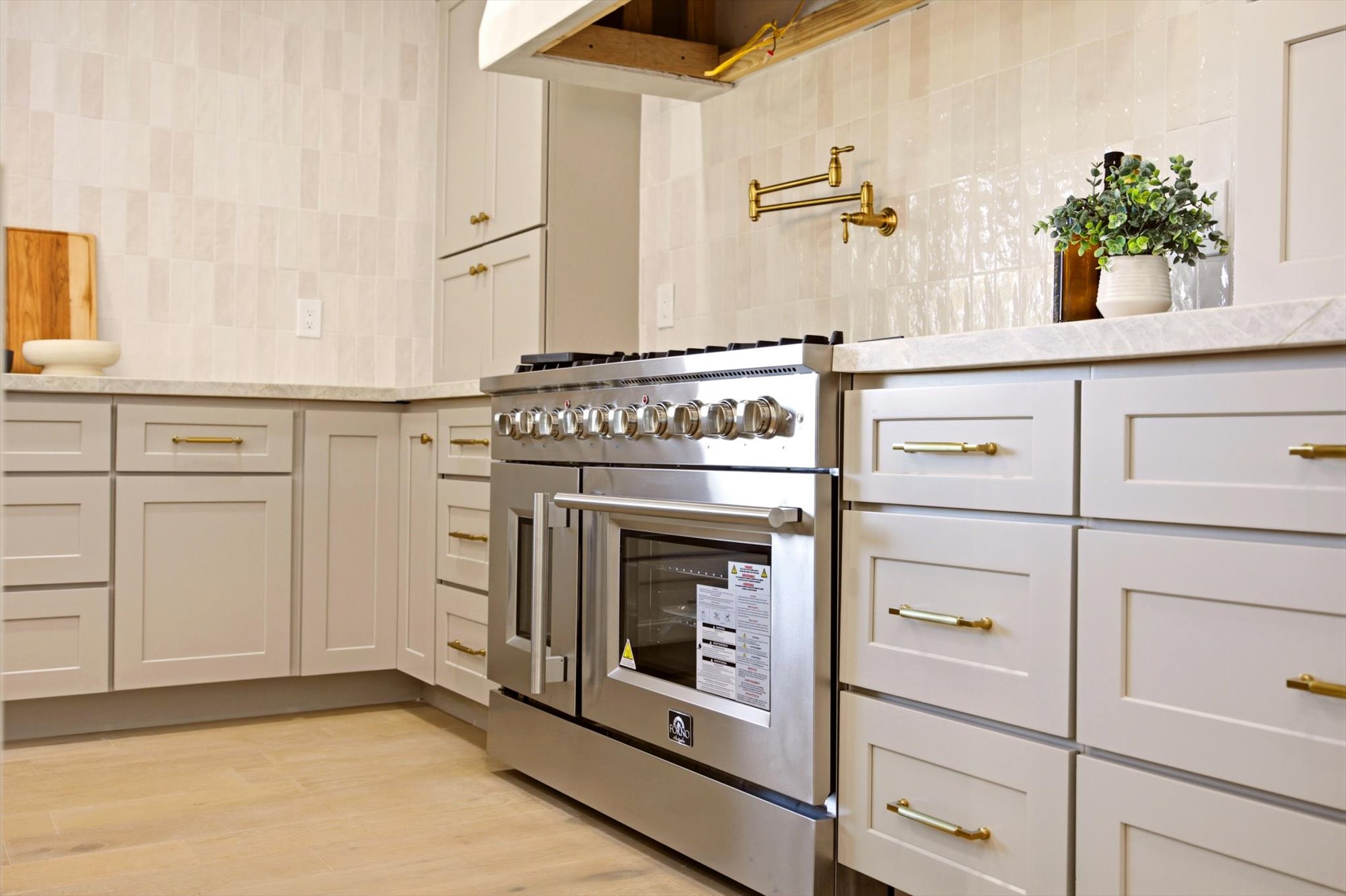 Gourmet kitchen featuring a stainless steel professional range, light-toned cabinetry with gold-finish hardware, light-colored countertops, a textured tile backsplash, and a wall-mounted gold-finish pot filler
