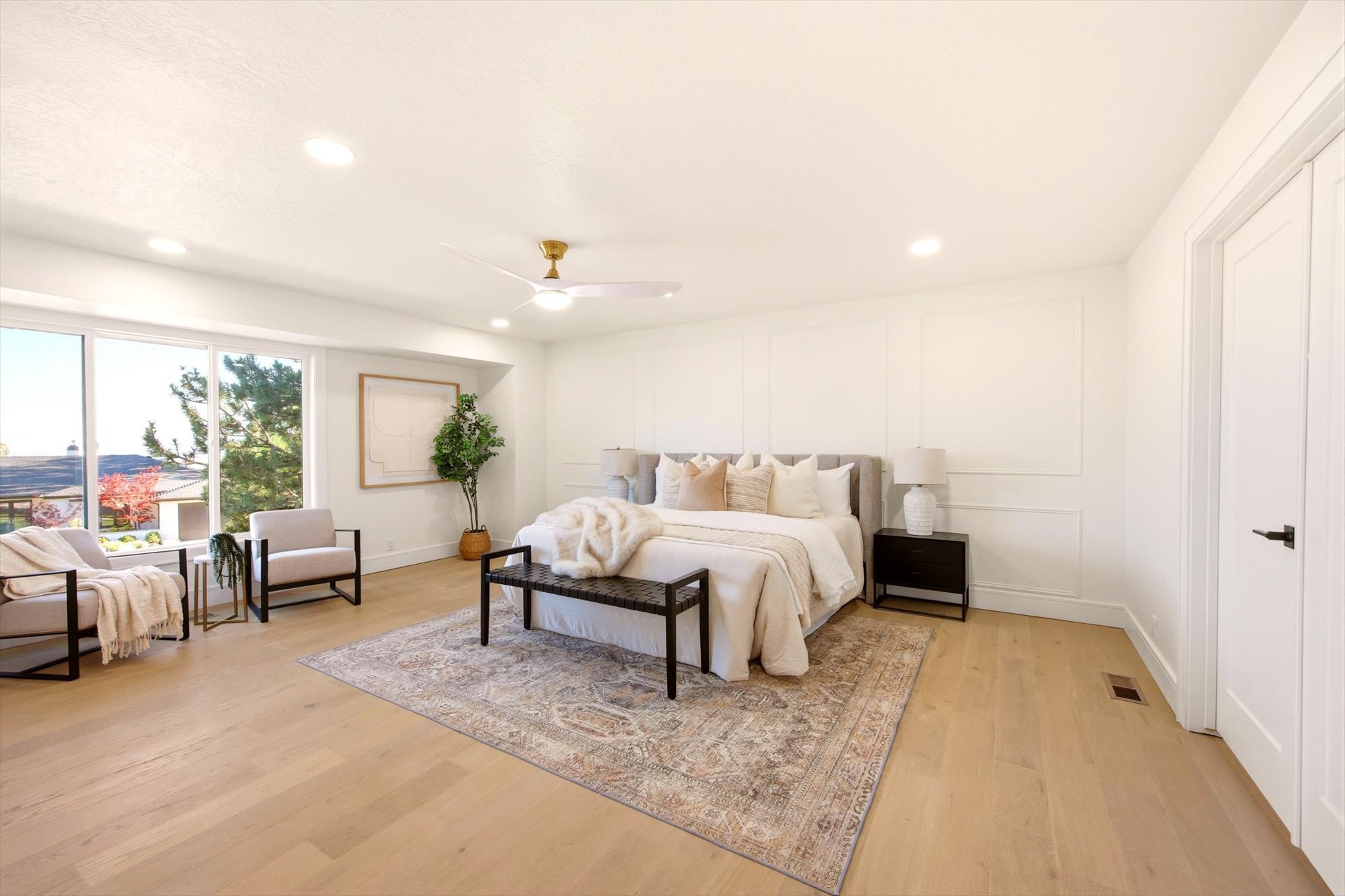 Spacious bedroom featuring light wood-finish flooring, a wall with decorative paneling, recessed lighting, and a ceiling fan with a gold-tone accent