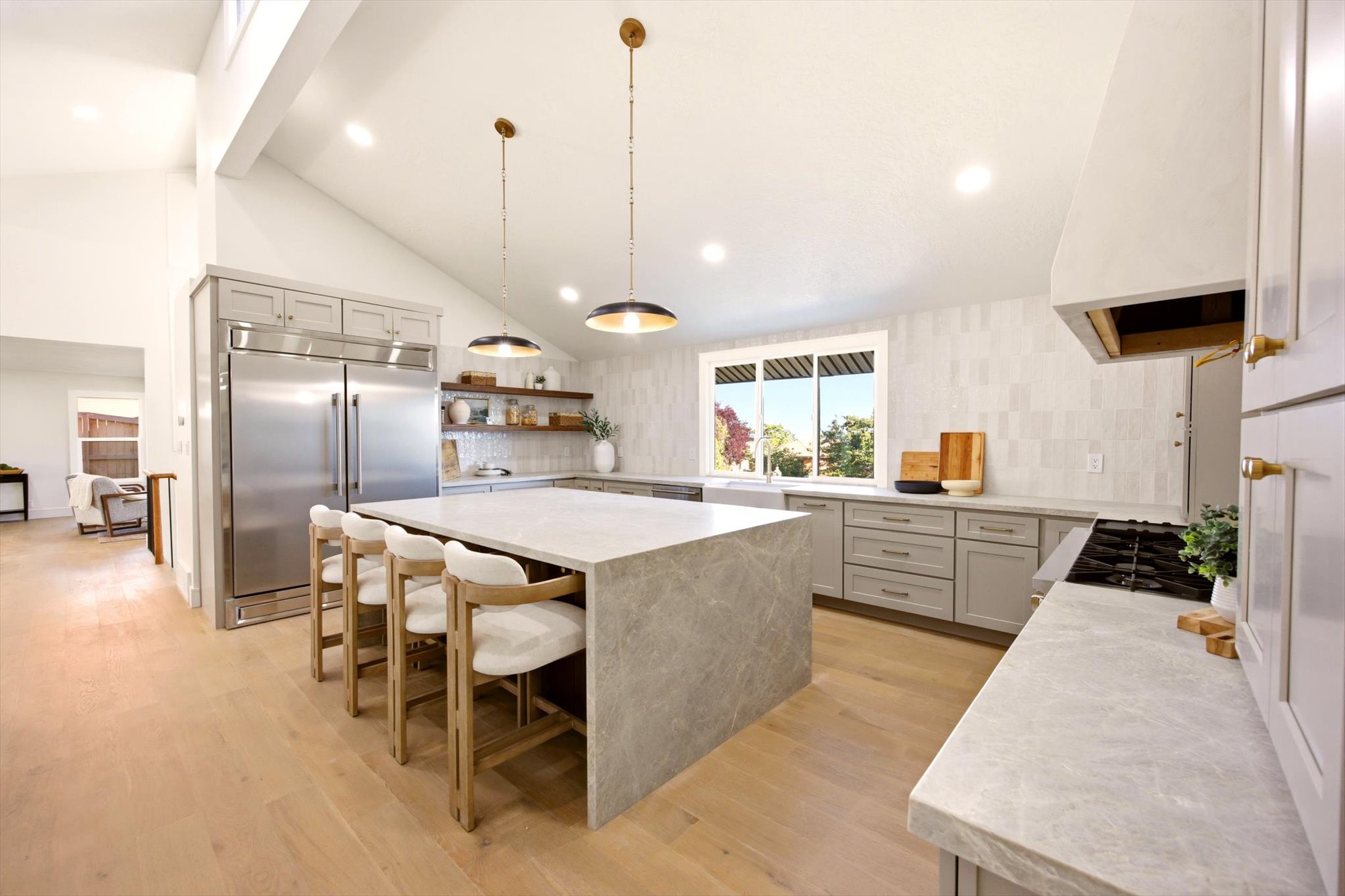 Spacious kitchen featuring a large central island with waterfall countertop, light wood-finish flooring, a vaulted ceiling, stainless steel refrigerator, and recessed lighting