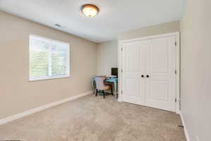 Neutral-toned room featuring a single window with blinds, light-colored carpeting, white baseboards, and a pair of white bifold closet doors with dark hardware