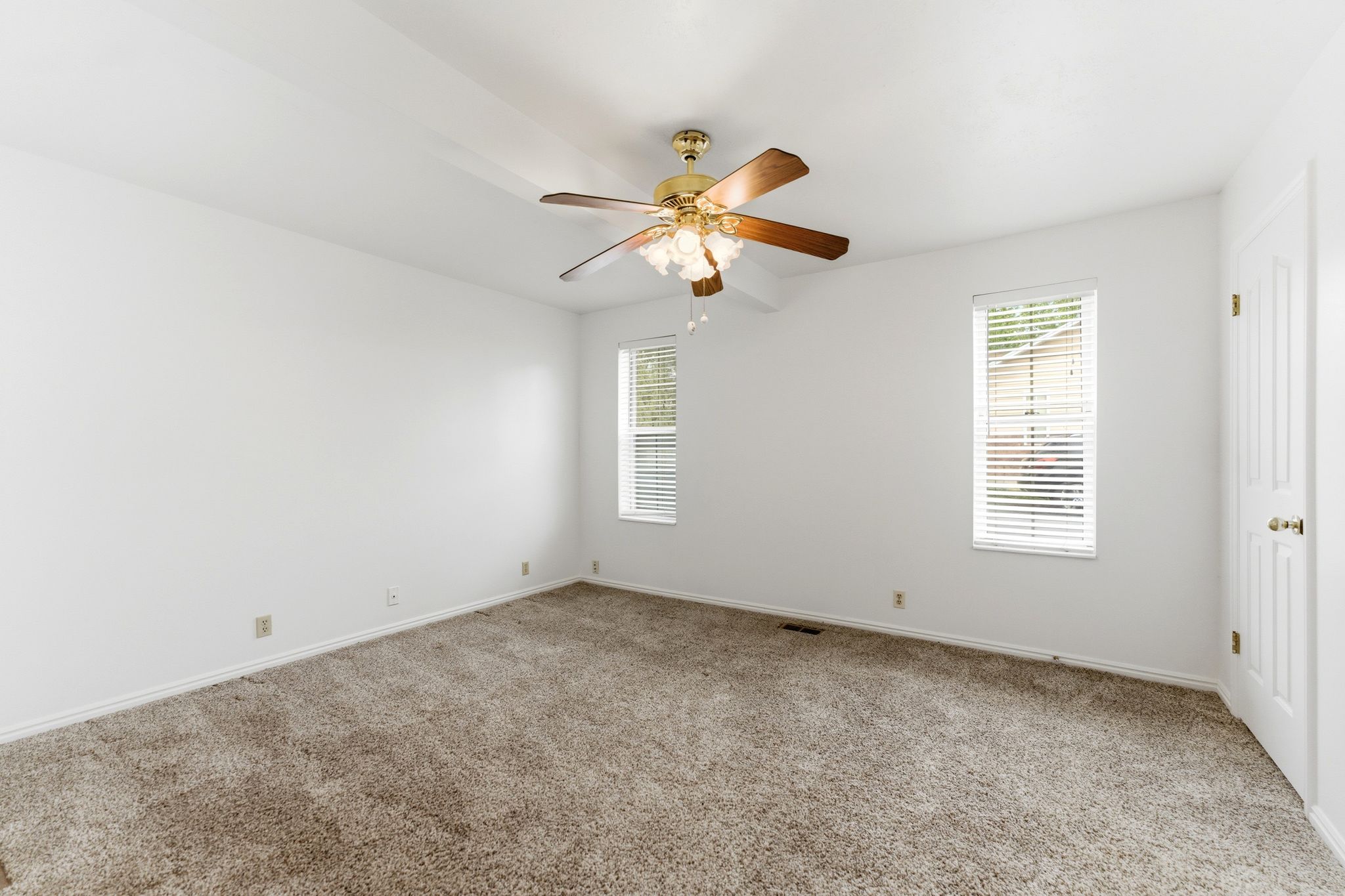 Spacious carpeted room featuring a ceiling fan with light fixture, two windows with blinds, and white walls