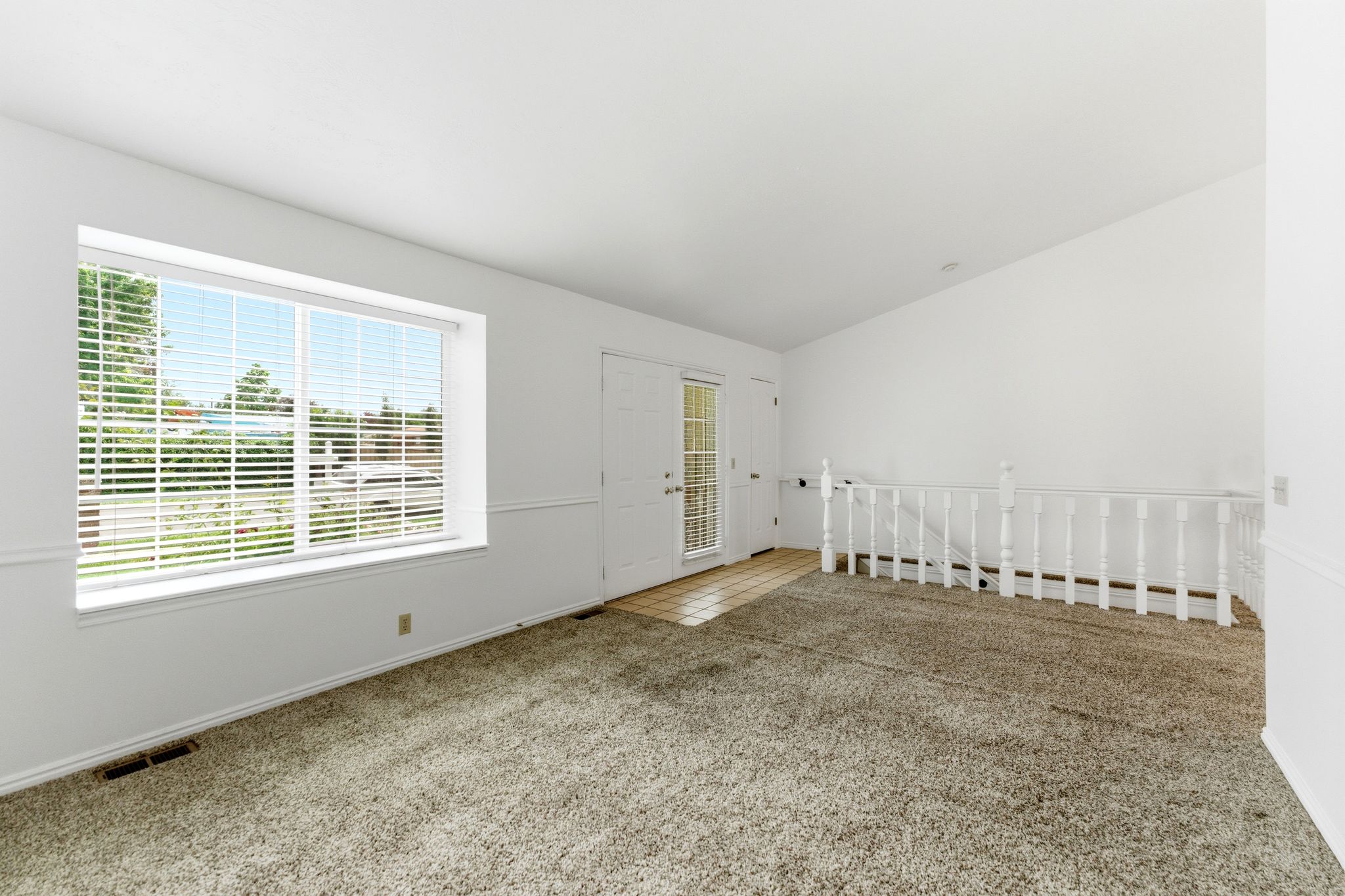 Spacious room featuring a large window with horizontal blinds, neutral carpet flooring, a white spindle railing, and a glass-paneled door