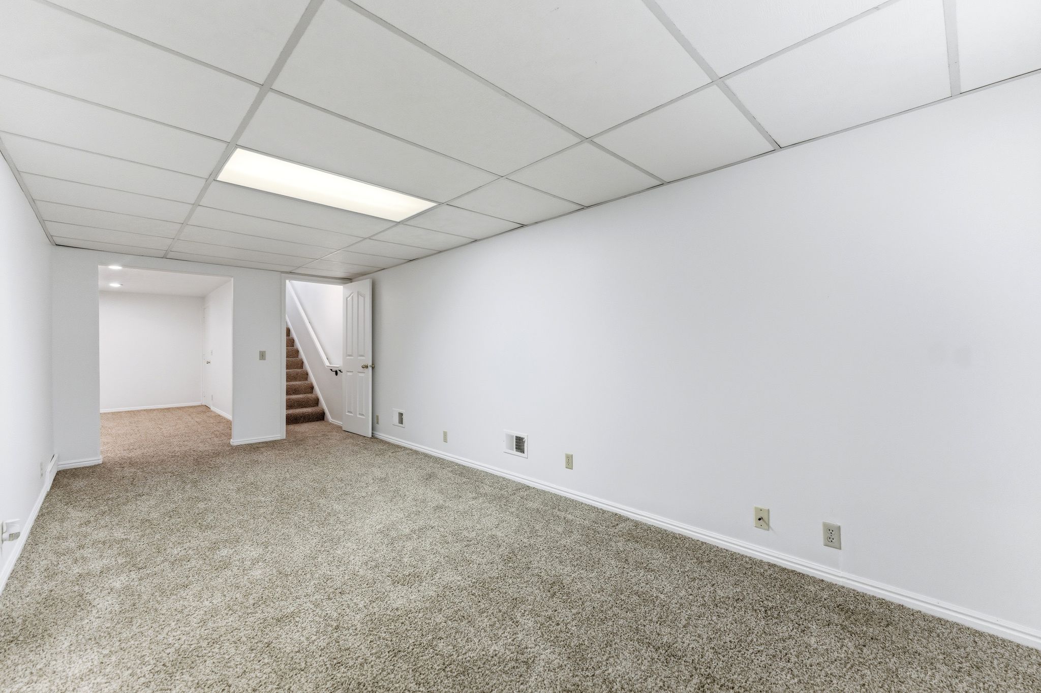 Spacious room featuring neutral carpeting, white walls, a drop ceiling with recessed lighting, and a visible staircase with a white door