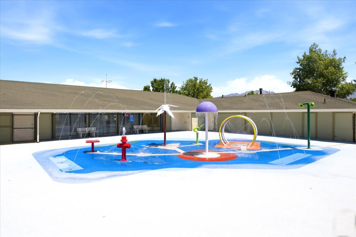 Community splash pad featuring multiple water features, a soft-surface play area, and perimeter buildings with covered walkways