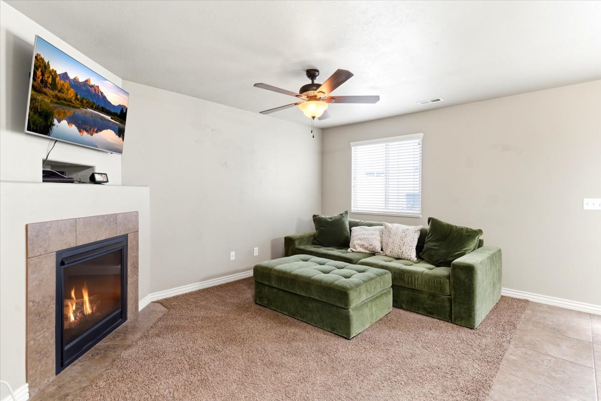 Living area featuring a gas fireplace with a tiled surround, a ceiling fan with integrated lighting, a window with blinds, and mixed flooring with carpet and tile sections