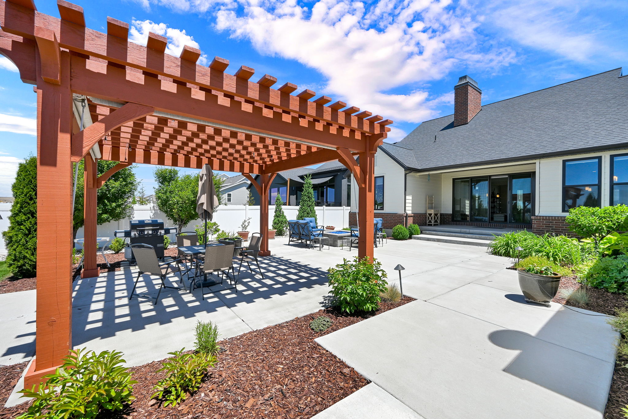 Spacious outdoor patio featuring a large wooden pergola, concrete hardscaping, mature landscaping, and a privacy fence