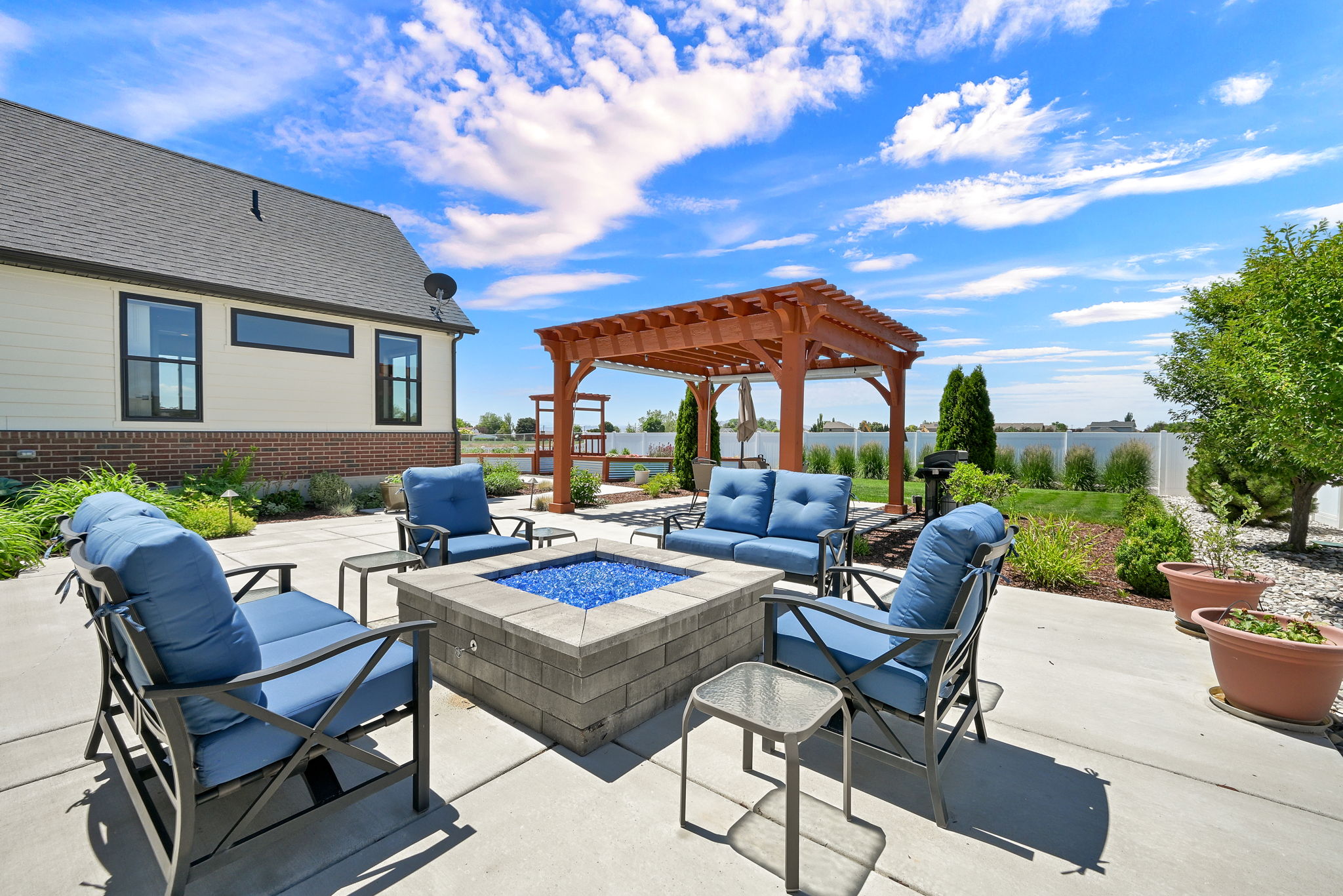 Expansive concrete patio featuring a gas fire pit with blue glass media, a wooden pergola, a manicured lawn, a privacy fence, and mature landscaping