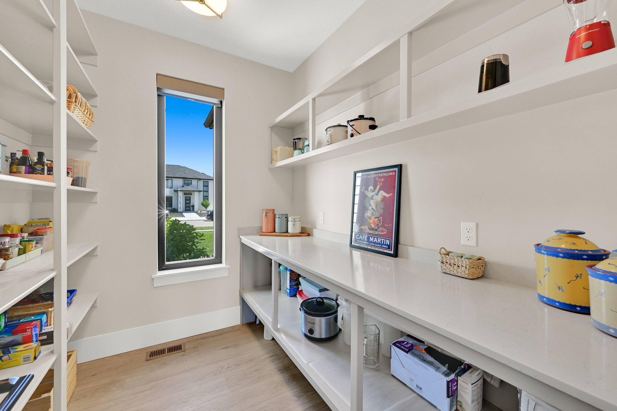 Spacious pantry featuring extensive open shelving, a large counter space with an under-counter shelf, wood-finish flooring, and a single vertical window providing natural light