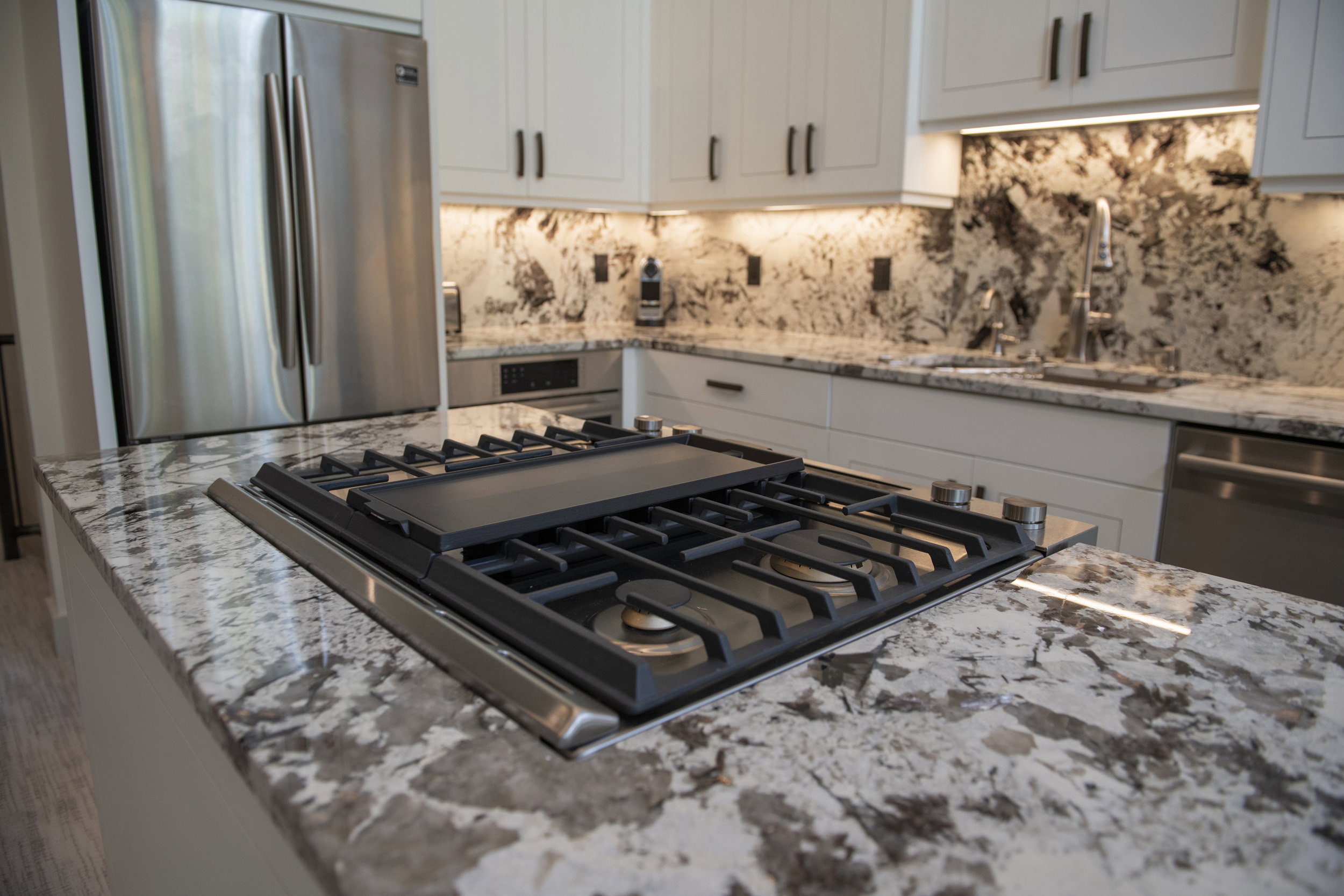 Gourmet kitchen featuring a built-in gas range with griddle, stainless steel refrigerator, white cabinetry with dark hardware, a full-height stone backsplash, and recessed under-cabinet lighting