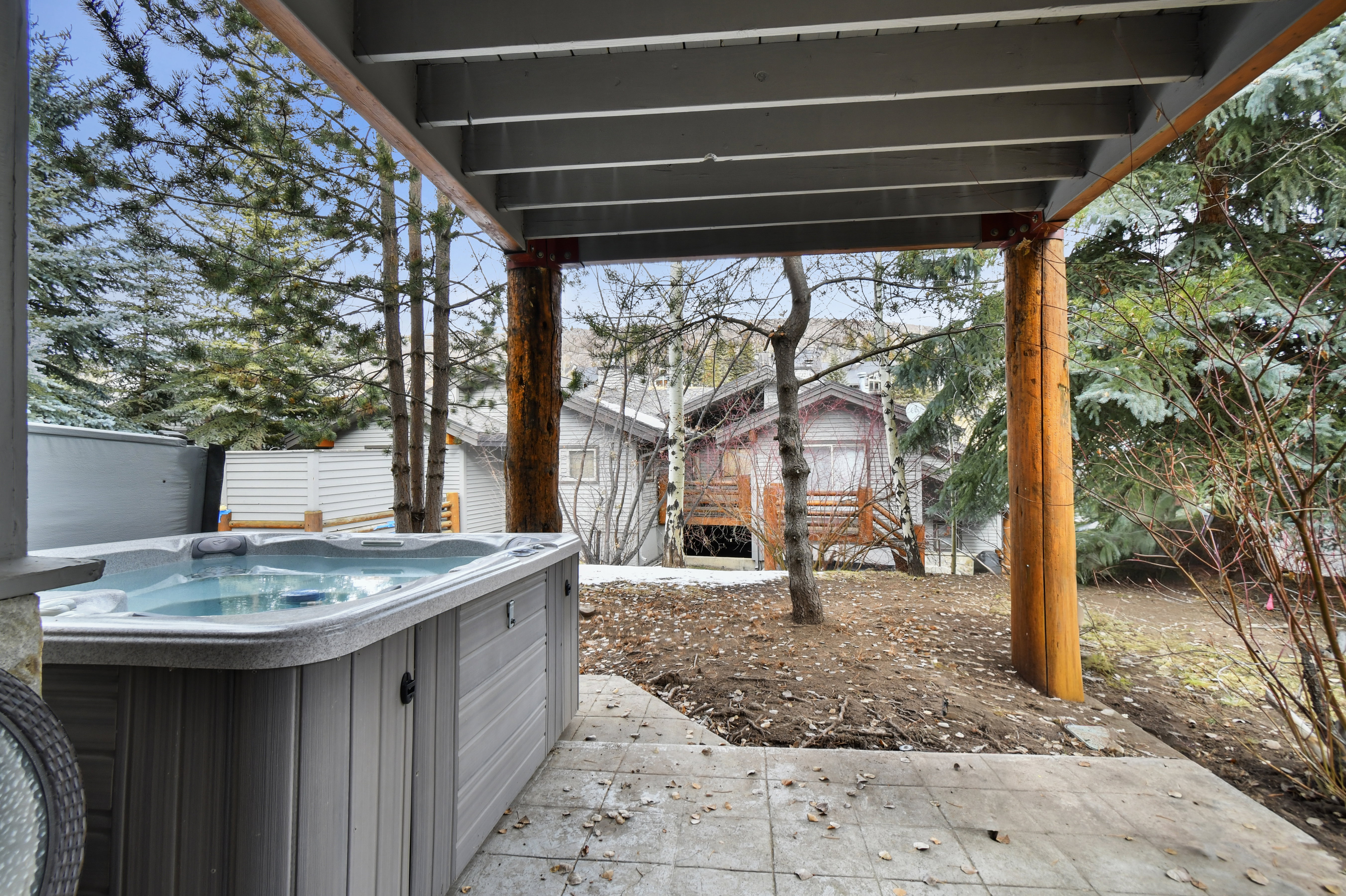 Paved patio area featuring a built-in spa, overhead deck structure with rustic wood posts, and mature evergreen trees