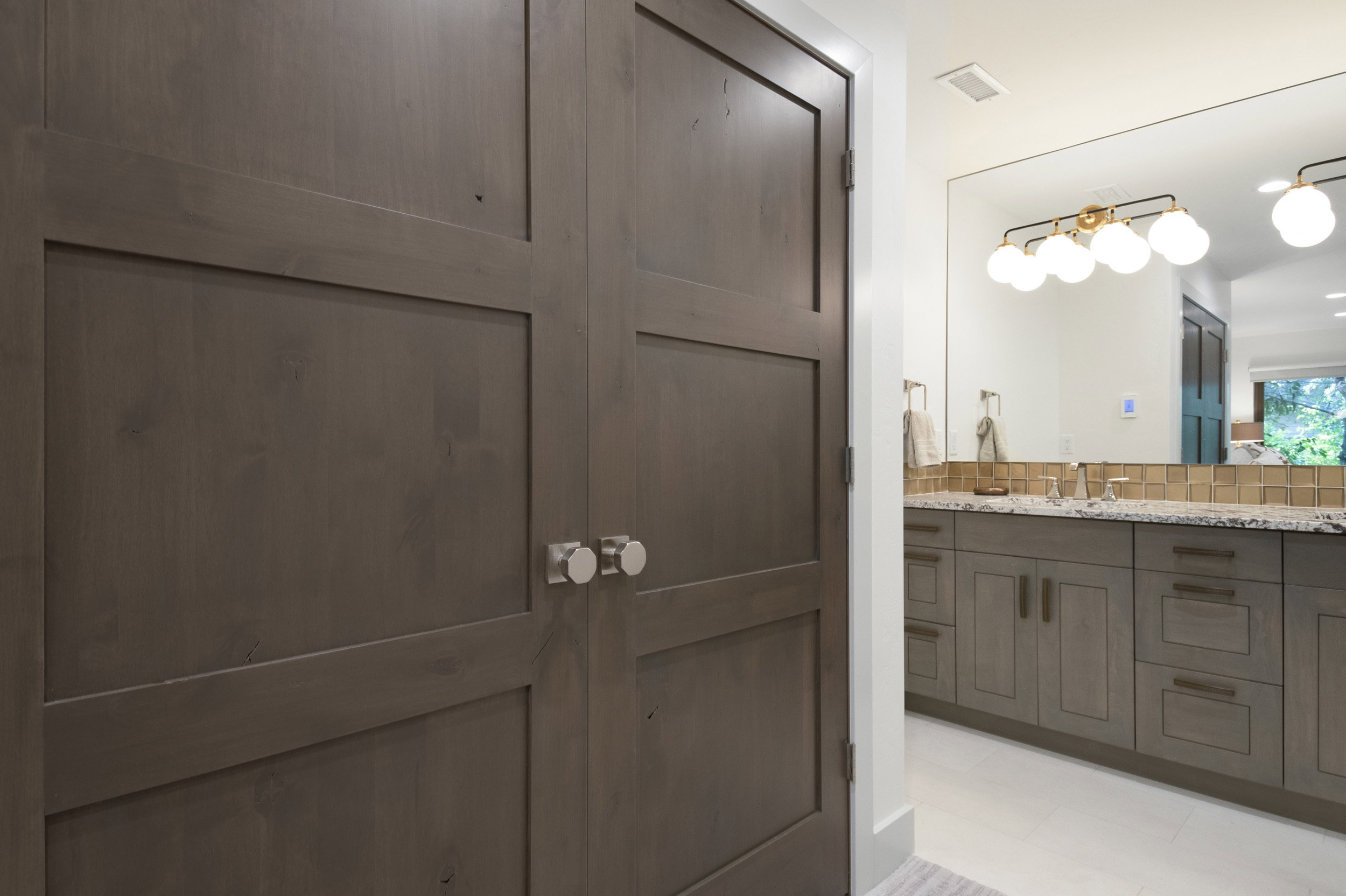 Double bi-fold doors with round brushed nickel hardware, light wood-finish vanity with drawer and cabinet storage, stone countertop, dual undermount sinks, and a large frameless mirror with a multi-bulb vanity light fixture
