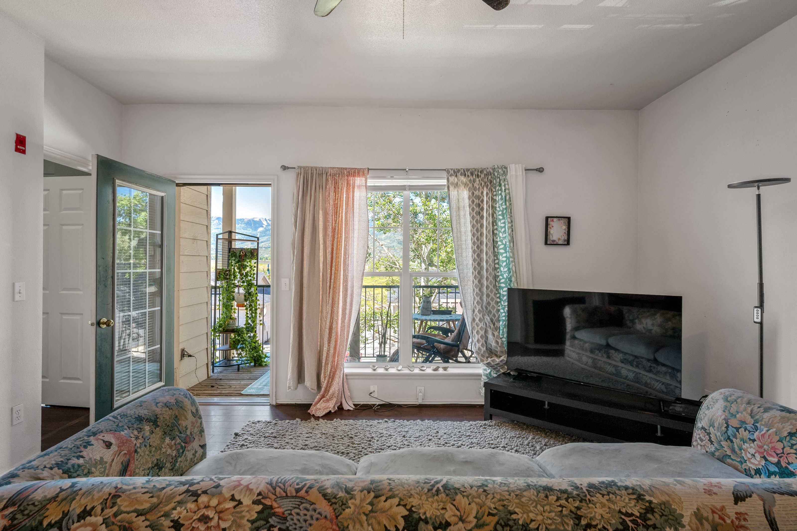 Living area with dark wood-finish flooring, white walls, a ceiling fan, and a glass-paneled door opening to a balcony