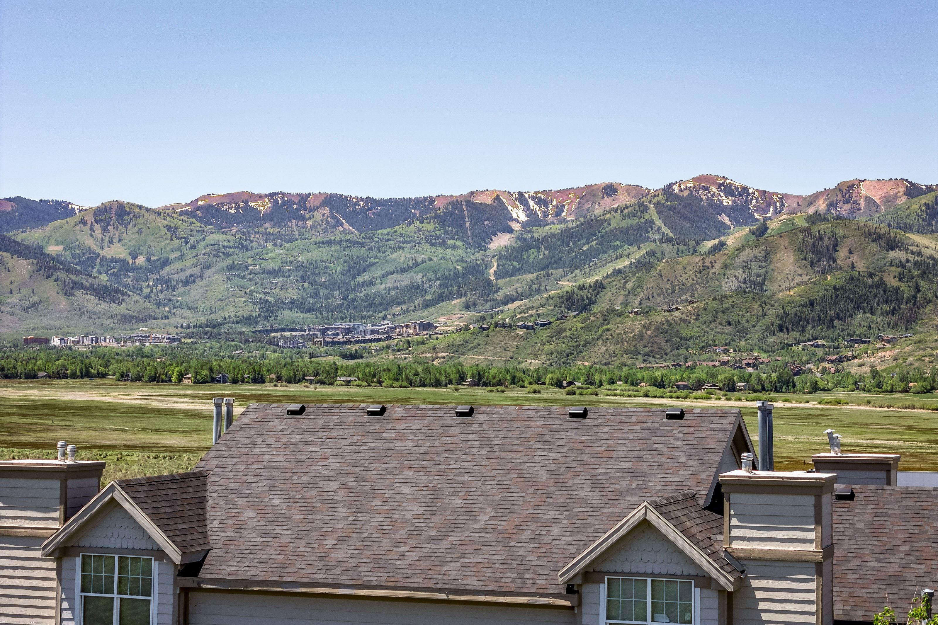 Mountain landscape with expansive green valleys and distant treeline, framed by a residential roofline featuring multiple chimneys and gabled dormers