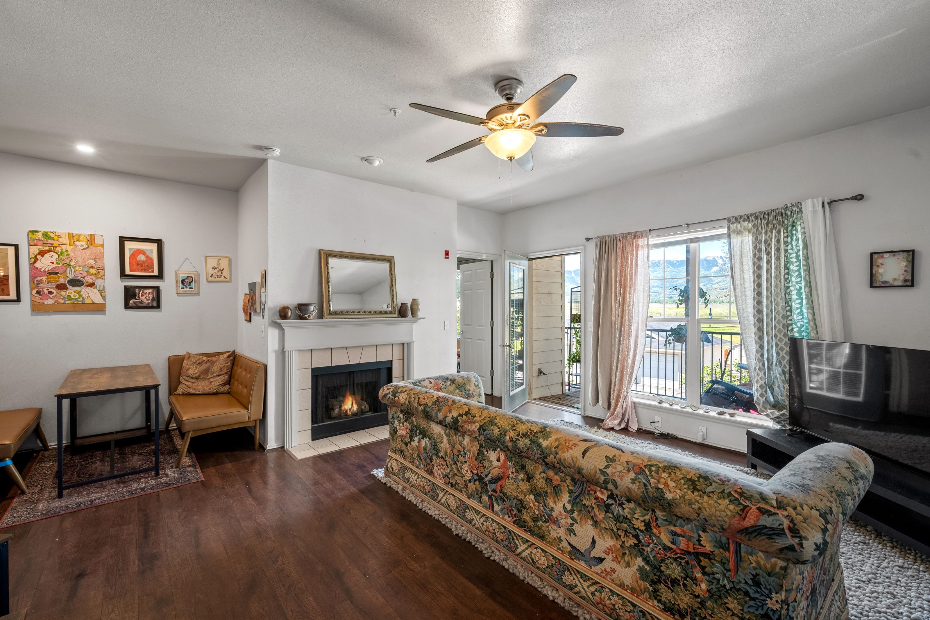 Living area featuring wood-finish flooring, a fireplace with a white mantel and tile surround, and a ceiling fan with integrated lighting
