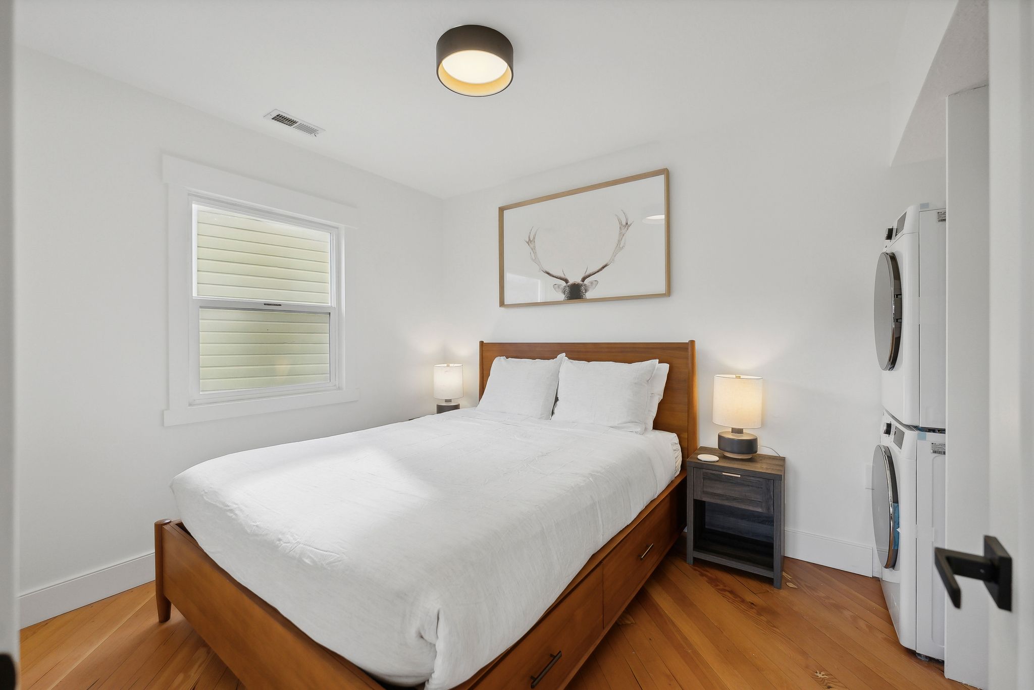 Bedroom featuring wood-finish flooring, a contemporary ceiling light fixture, a window with horizontal blinds, and a stacked washer and dryer unit