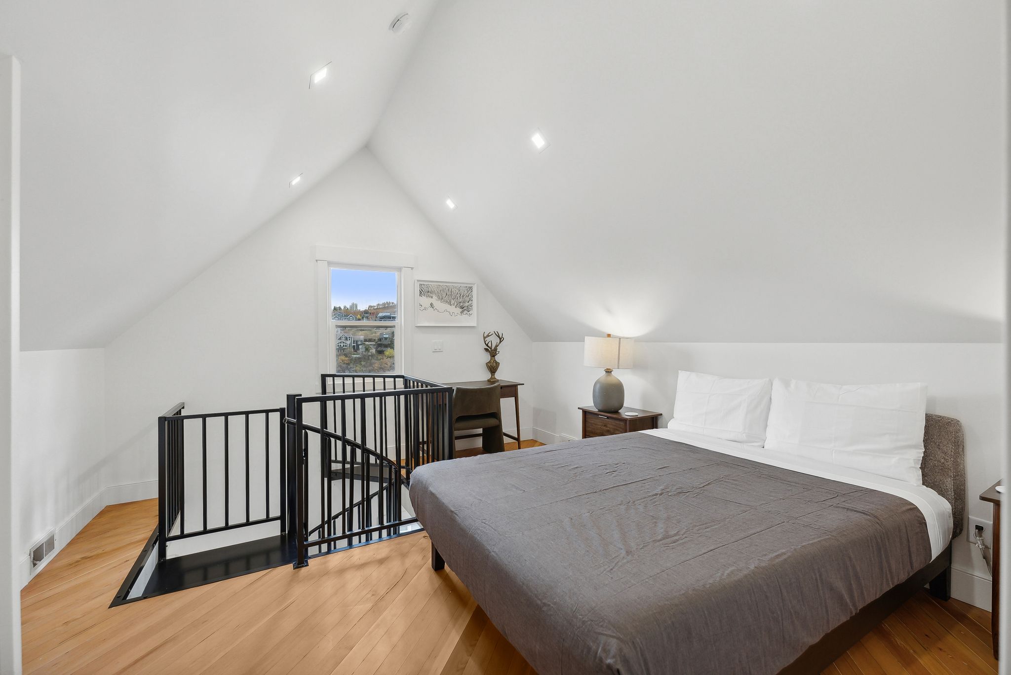 Attic bedroom featuring pitched ceilings, recessed lighting, wood-finish flooring, a window offering an outdoor perspective, and a black wrought iron railing enclosing the stairwell opening