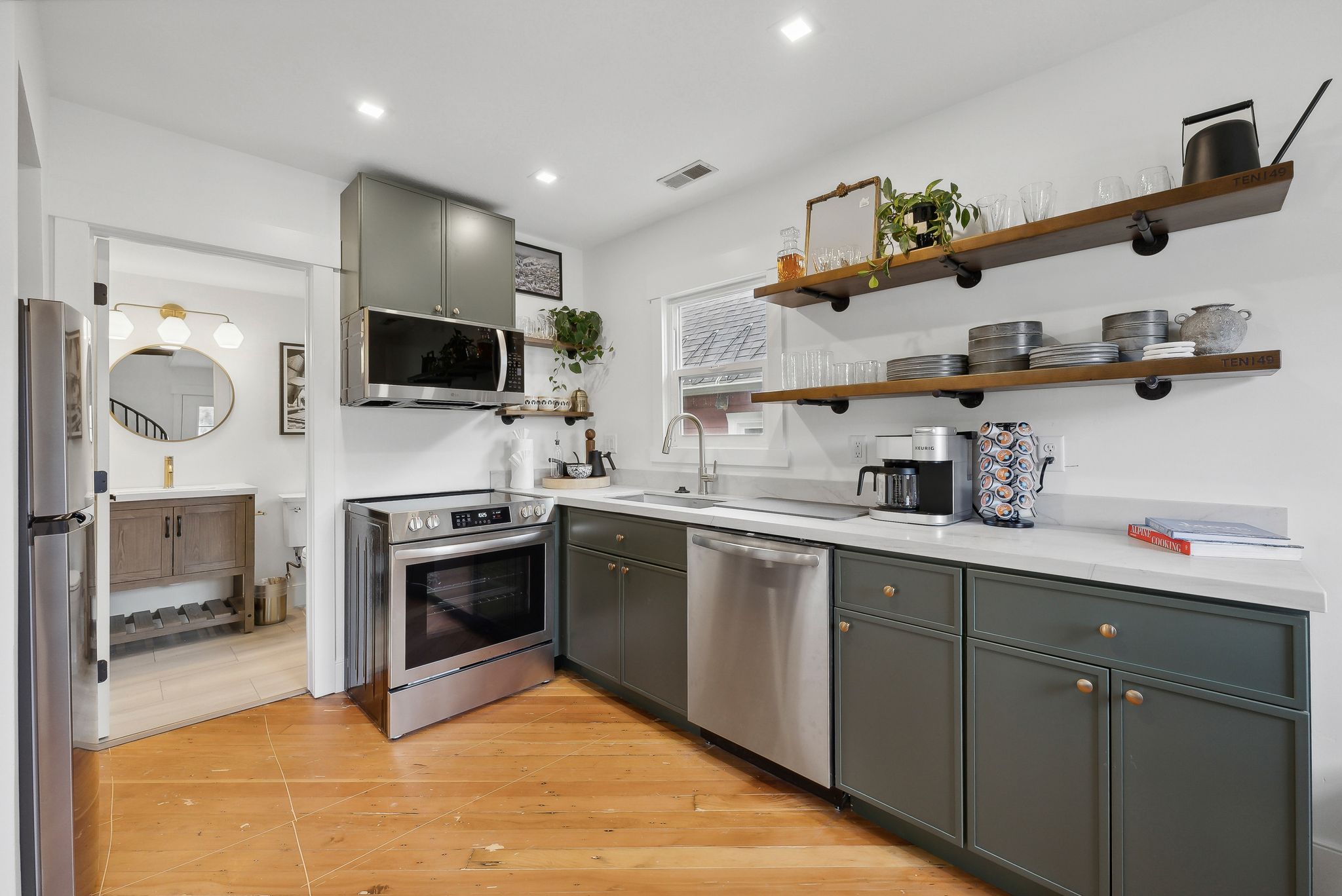 Kitchen featuring wood-finish flooring, dark green cabinetry with brass-toned hardware, stainless steel appliances, open shelving, and light-toned countertops
