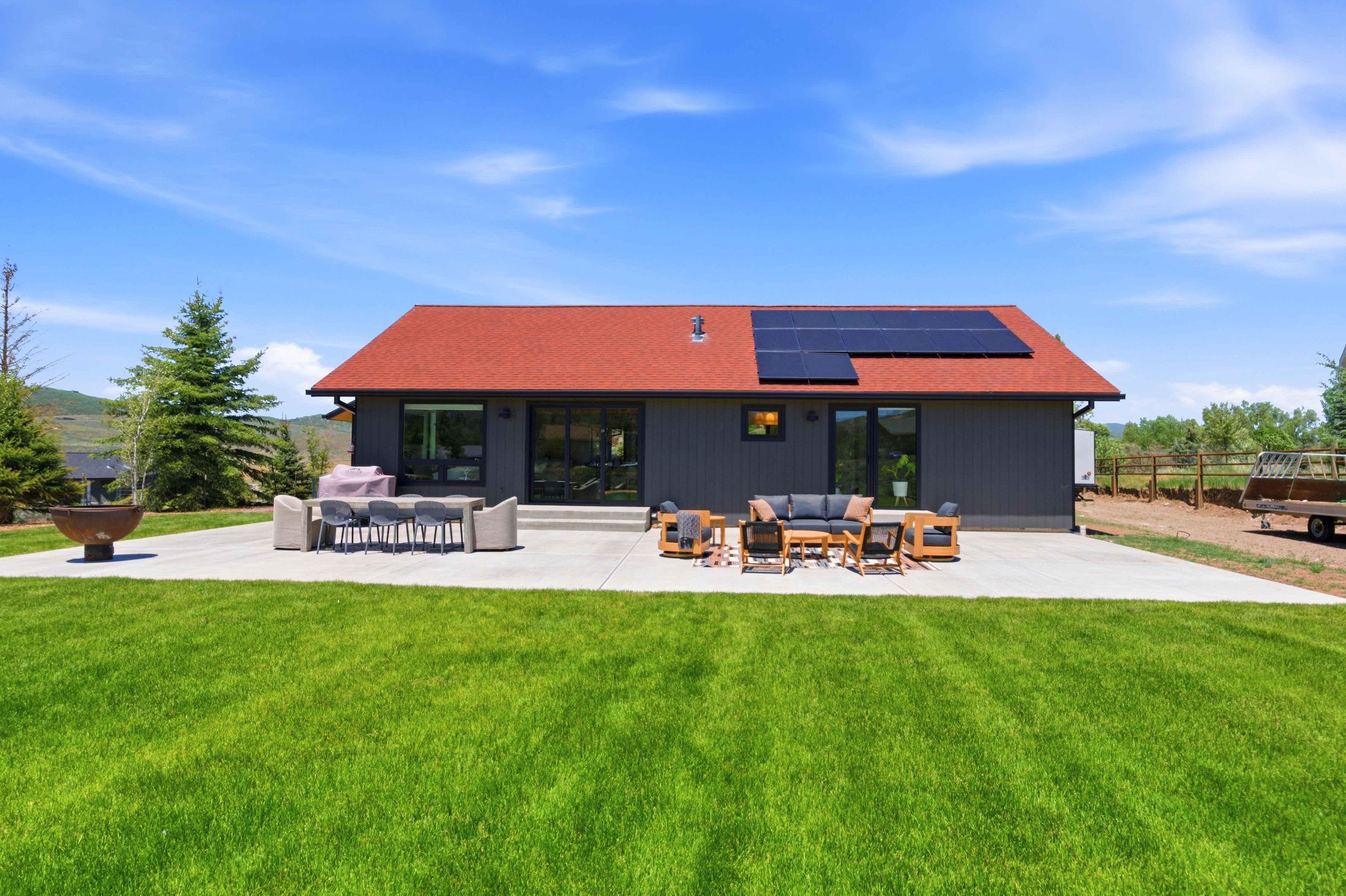 Expansive green lawn with a concrete patio featuring a modern house with dark siding, a red roof, and roof-mounted solar panels