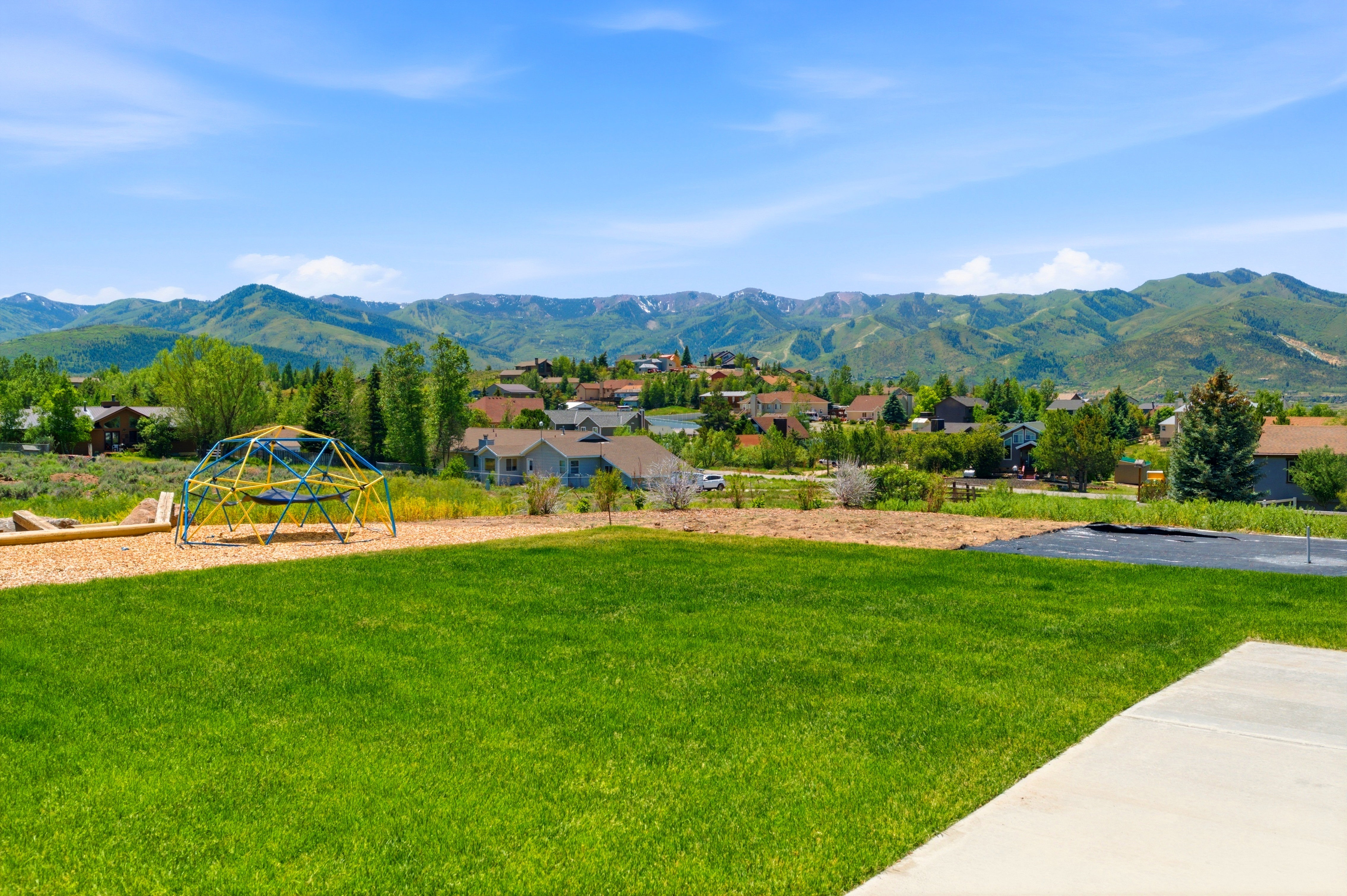 Expansive green lawn with a concrete patio, bordered by a wood chip play area featuring a climbing dome