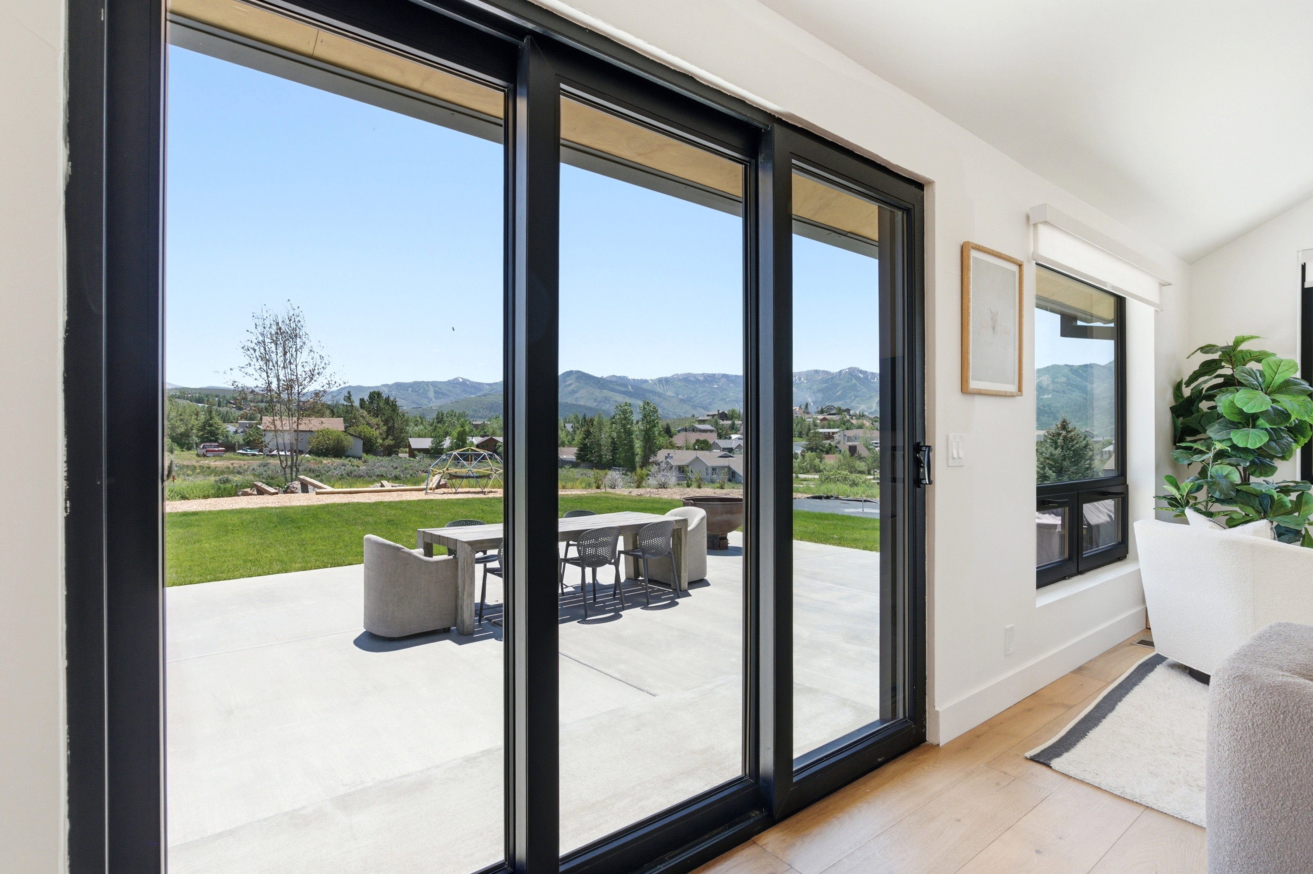 Black-framed sliding glass doors opening to a concrete patio with a fire pit feature, surrounded by a green lawn and distant mountain scenery