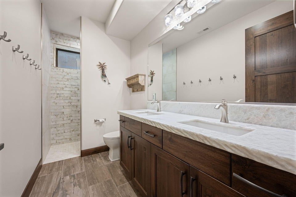 Contemporary bathroom featuring a dual vanity with undermount sinks, stone countertops, wood-finish cabinetry, wood-finish flooring, and a walk-in shower with brick-patterned tile and a fixed window