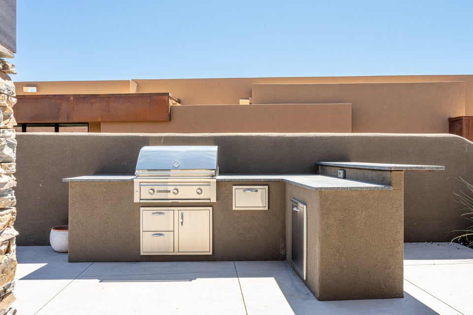 Outdoor L-shaped kitchen featuring a built-in stainless steel grill, integrated storage drawers, a compact refrigerator, and a granite countertop