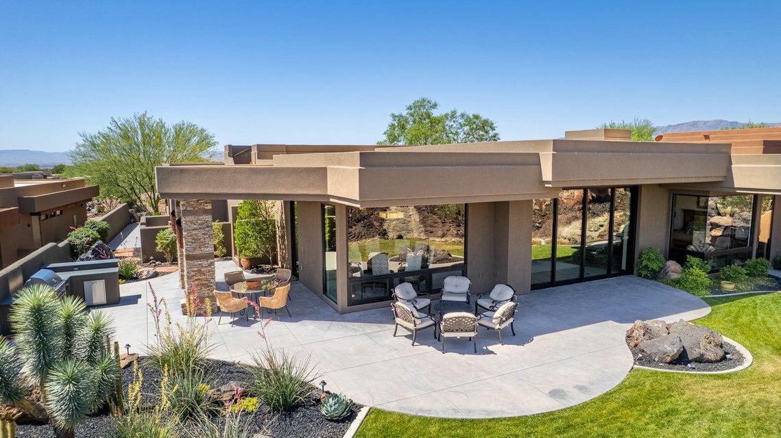Expansive concrete patio area with a built-in outdoor kitchen, stone accent pillars, full-height glass panels, and surrounding desert-inspired landscaping with a manicured lawn