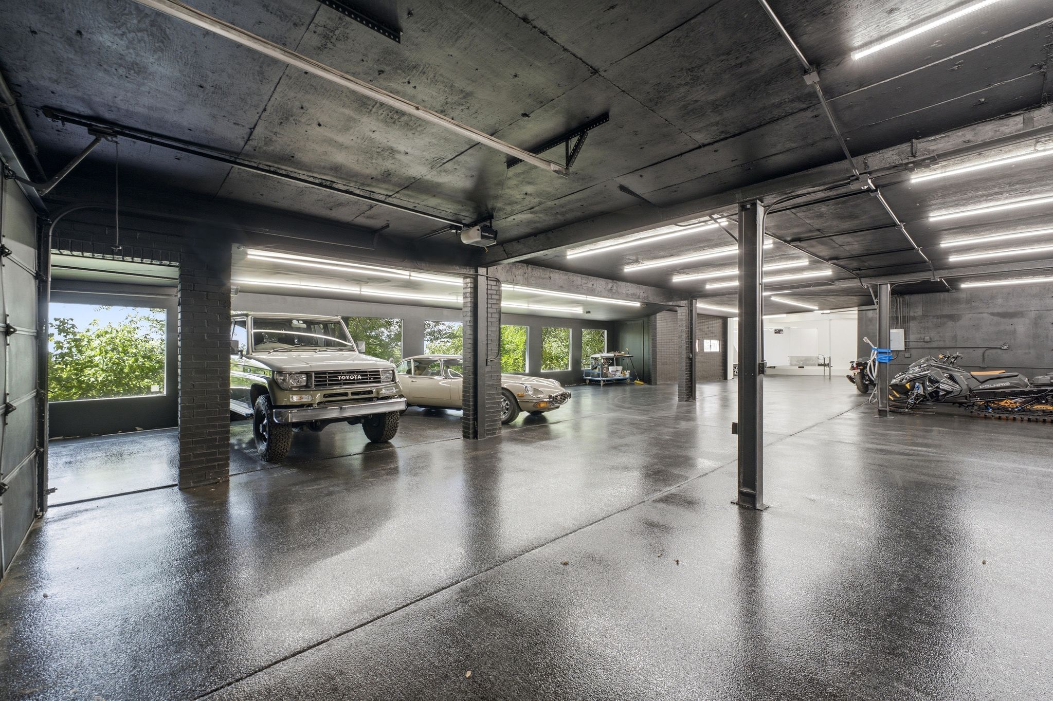 Spacious garage featuring a glossy epoxy floor, exposed concrete ceiling with strip lighting, multiple support columns, and an open garage door providing natural light