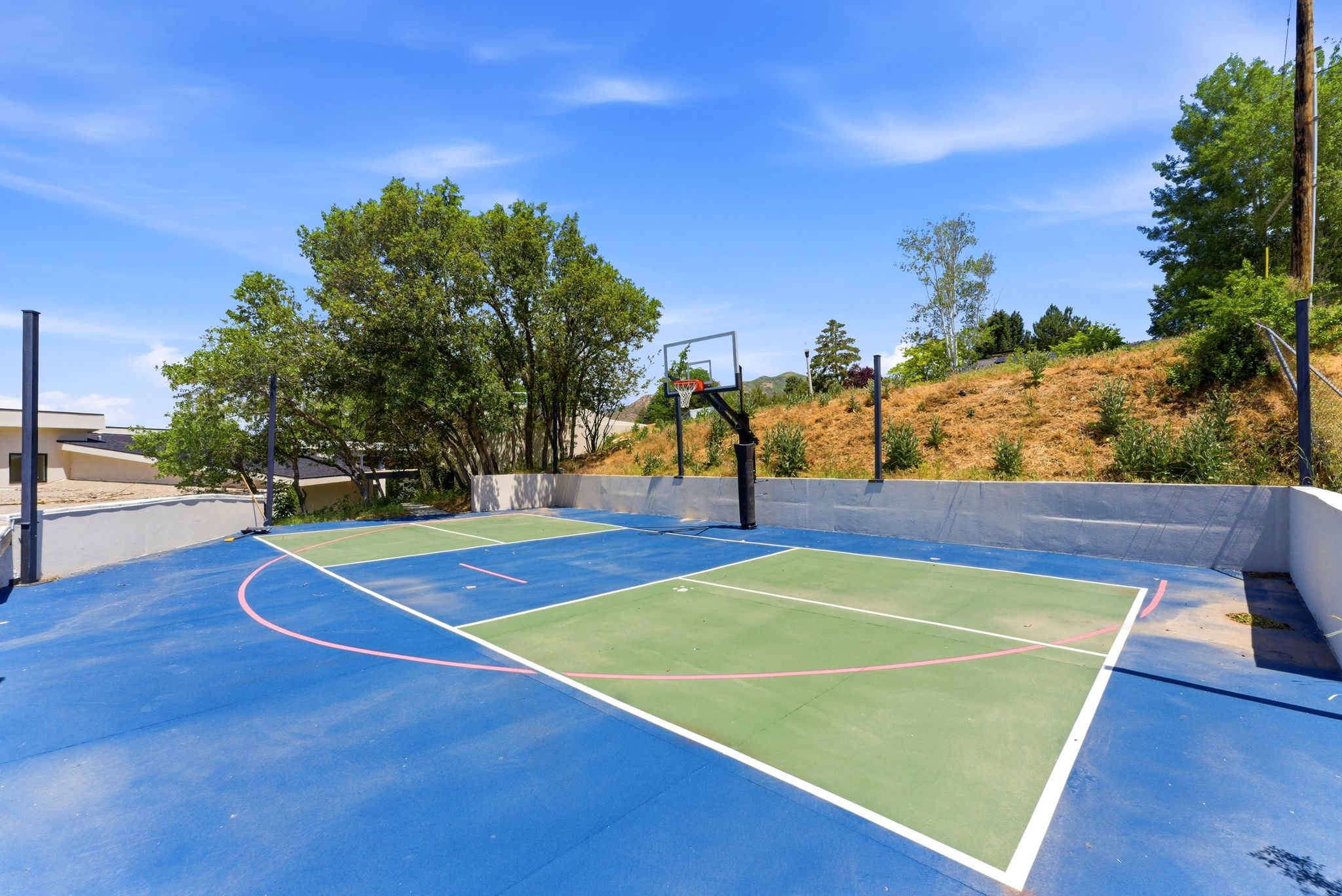 Dedicated outdoor sports court featuring a vibrant blue and green surface with white and pink boundary lines, a basketball hoop, and surrounding privacy walls