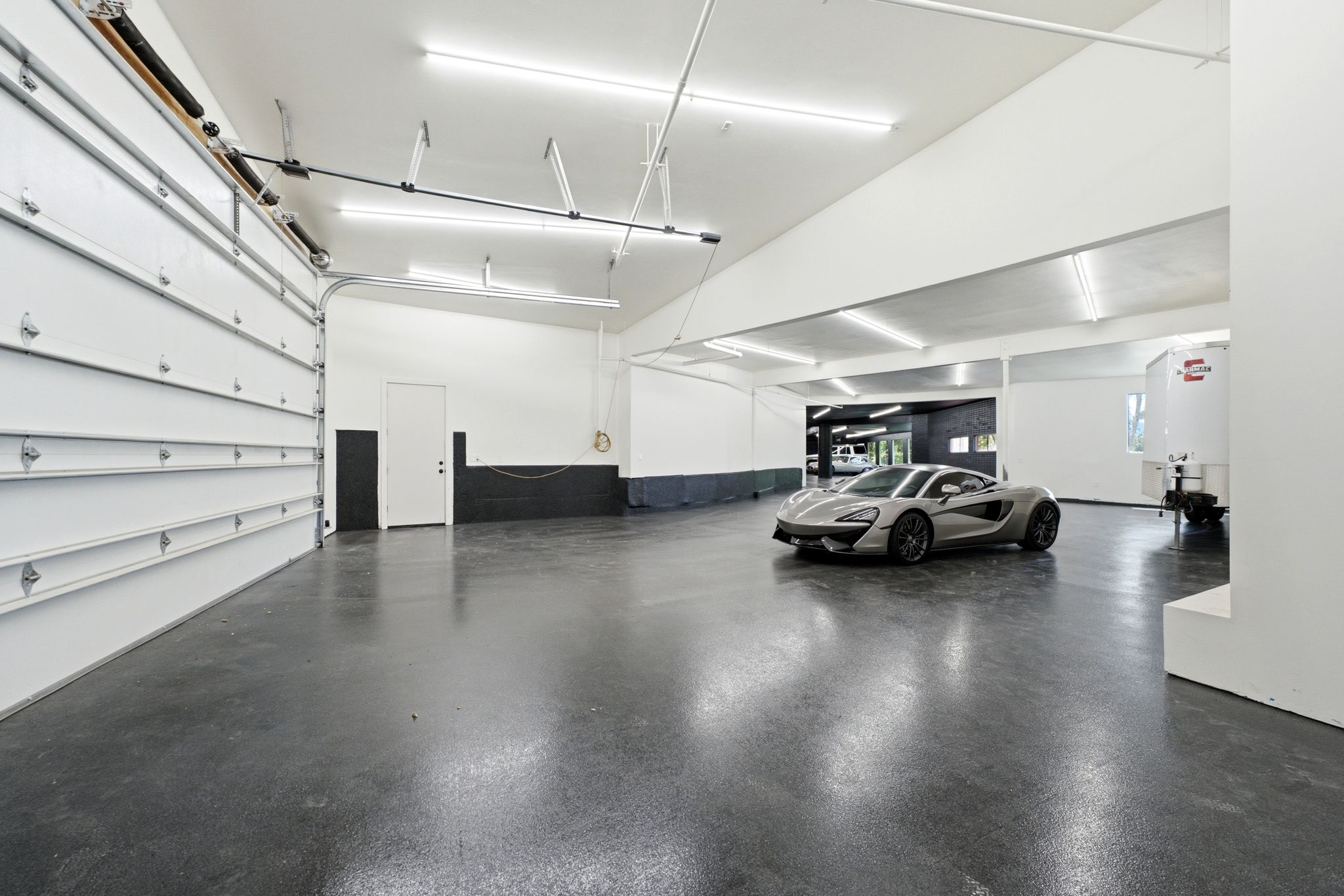 Spacious garage featuring a large roll-up door, high ceilings, polished concrete flooring, and ample fluorescent lighting