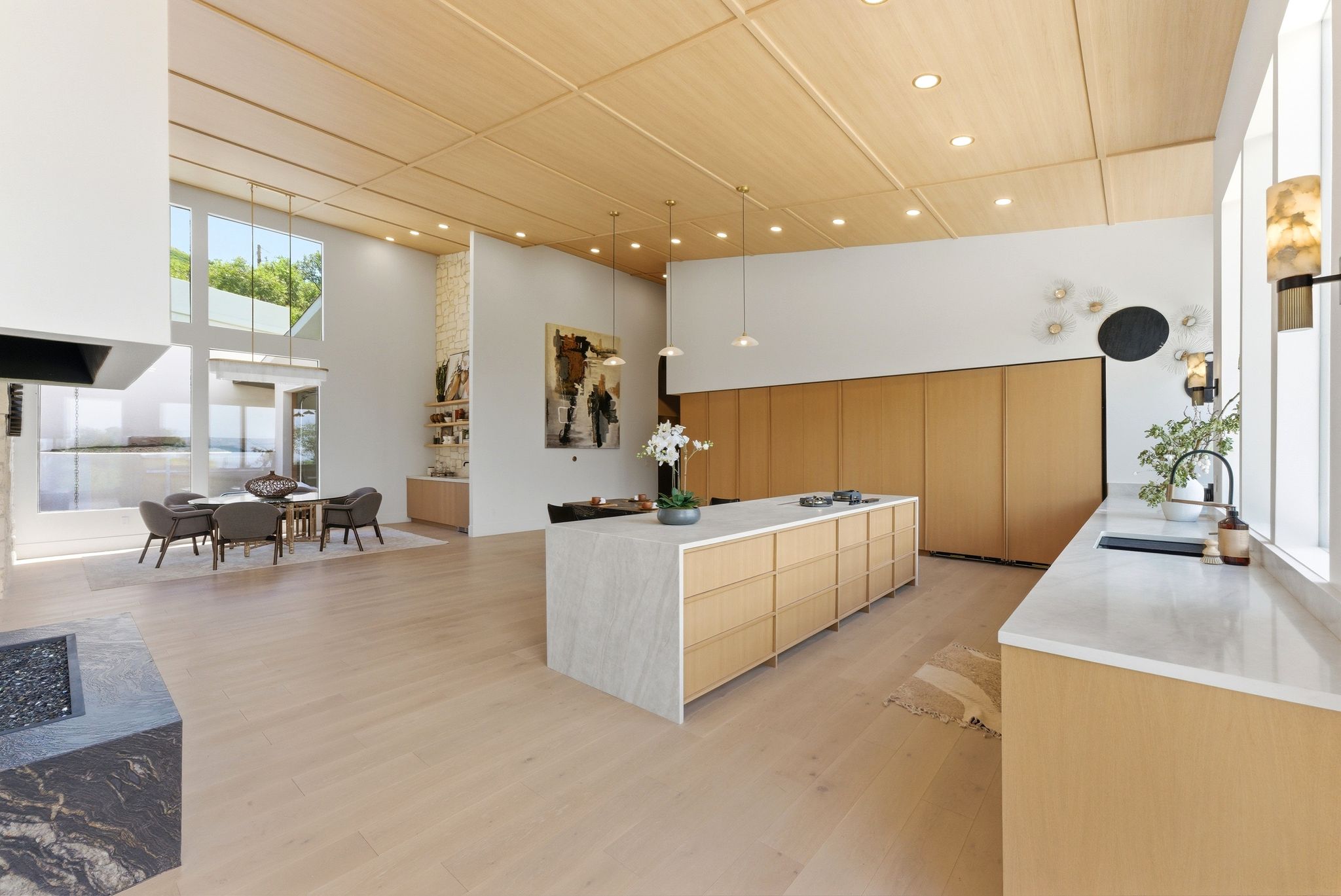 Open-concept kitchen and dining area featuring a large center island with integrated storage, wood-finish flooring, a light wood-paneled ceiling, a contemporary fireplace with a dark stone surround, and tall windows providing abundant natural light