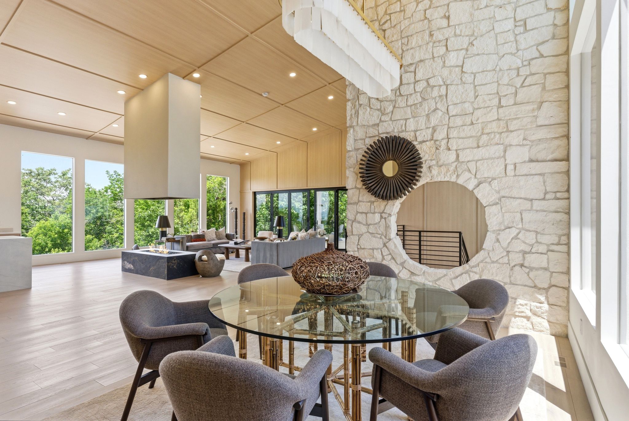 Expansive living area with a stacked stone accent wall featuring a circular opening, a floating ceiling with recessed lighting, large windows, and a central fireplace with a dark stone hearth