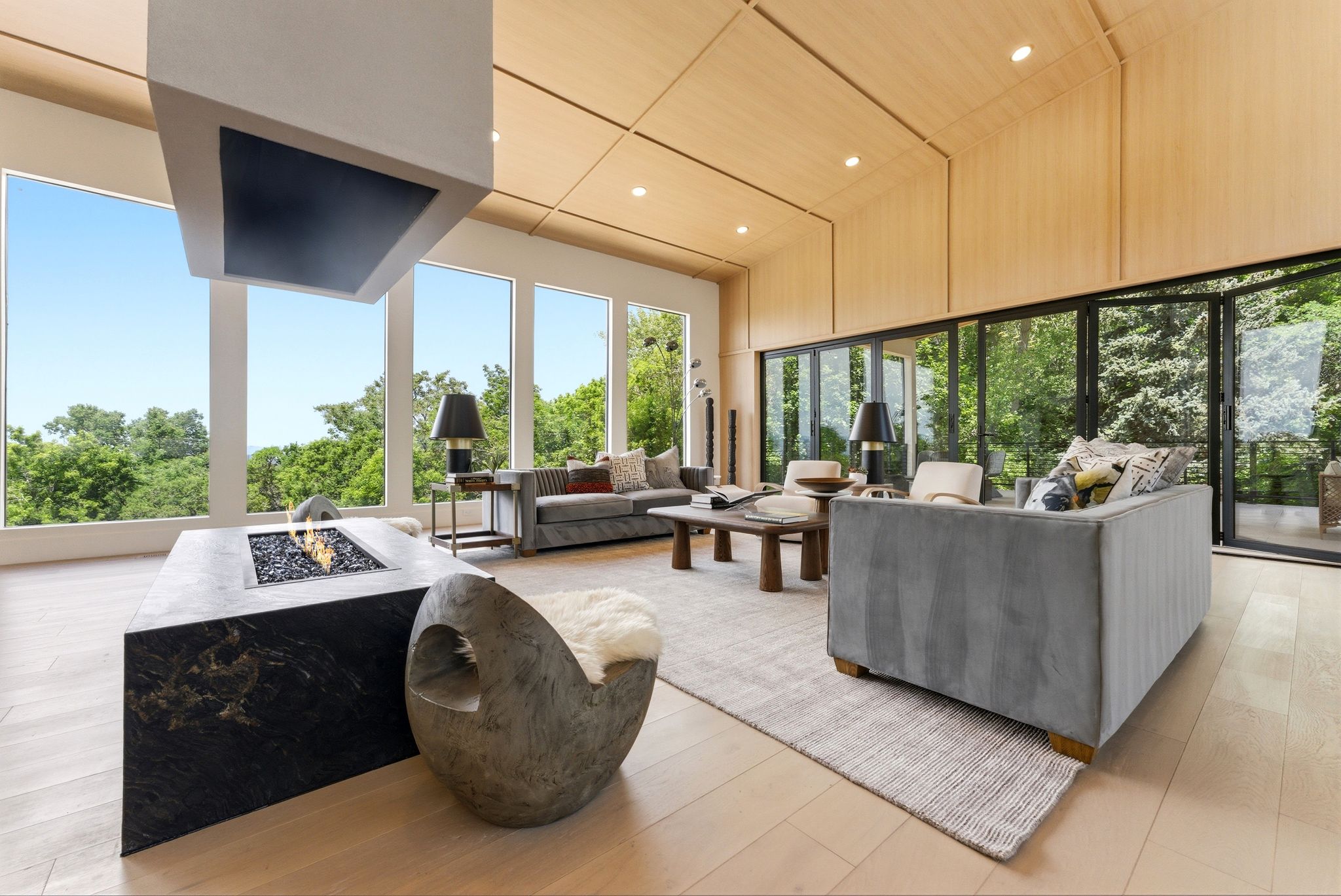 Expansive living area featuring a floating fireplace with a stone base, wood-finish flooring, a vaulted ceiling with integrated lighting, a wall of floor-to-ceiling windows, and multi-panel glass doors