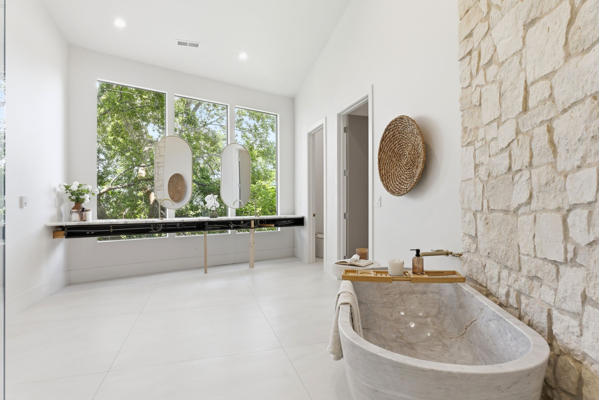 Spacious bathroom featuring a freestanding stone soaking tub, a full-height stone accent wall, and expansive windows