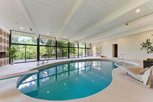 Indoor pool with integrated spa, full-height windows, a decorative accent wall, and a wood-paneled vaulted ceiling