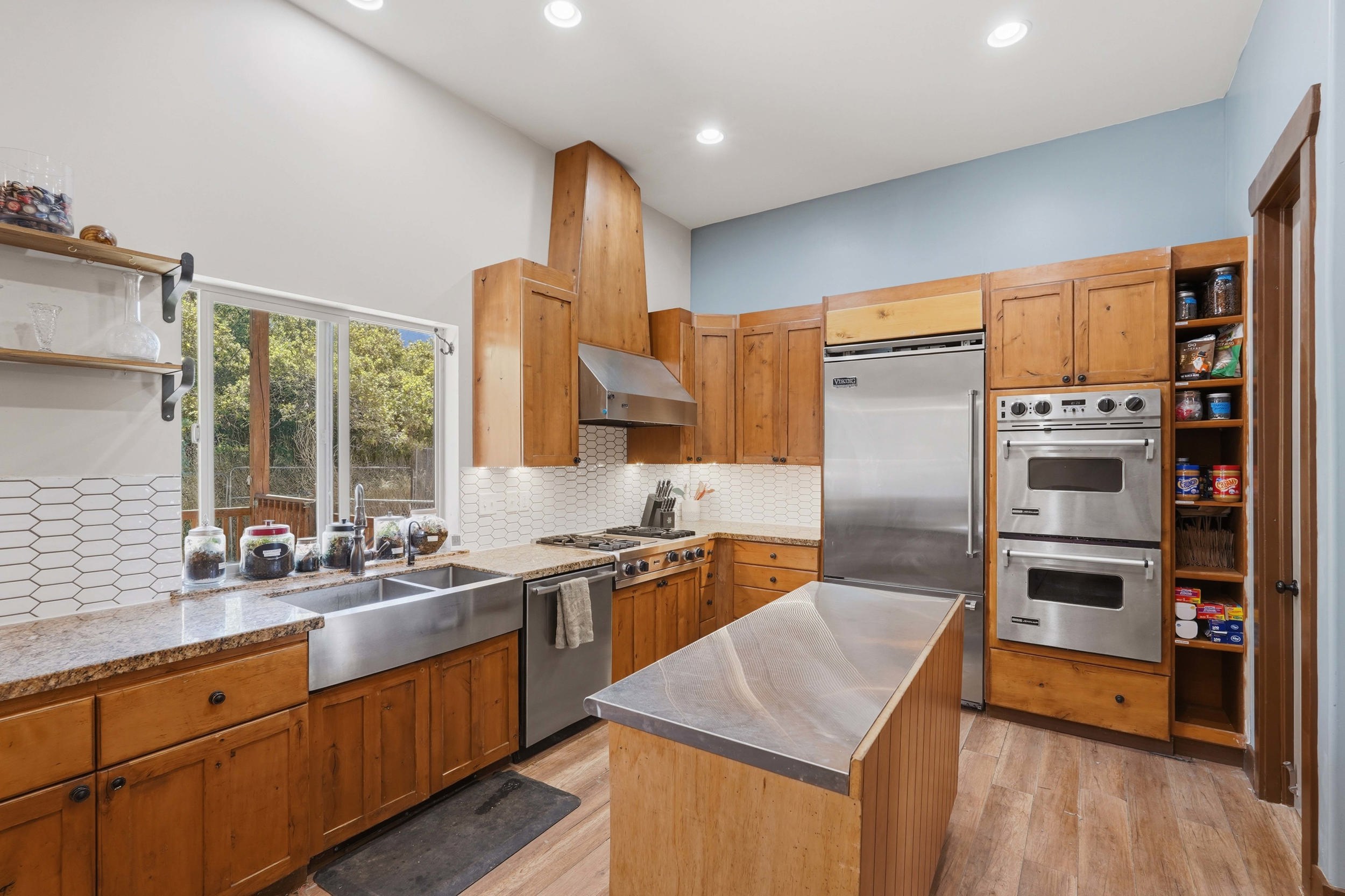 Kitchen featuring wood-finish cabinetry, stainless steel appliances, a large window, a farmhouse sink, and a tiled backsplash