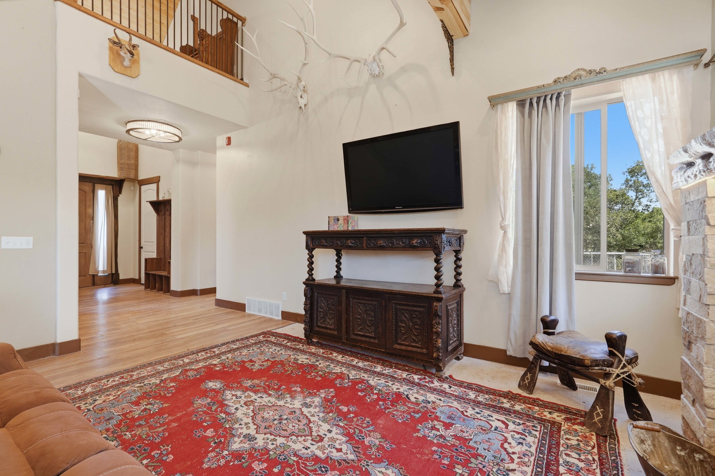 Two-story living area featuring a wood-finish flooring entryway, an upper-level wood railing, a stone-faced fireplace, and a window with exterior tree views