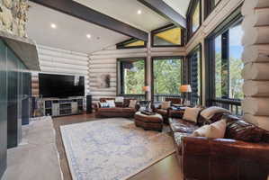 Vaulted ceilings with exposed beams, wood-finish flooring, a stone-clad fireplace, and expansive windows
